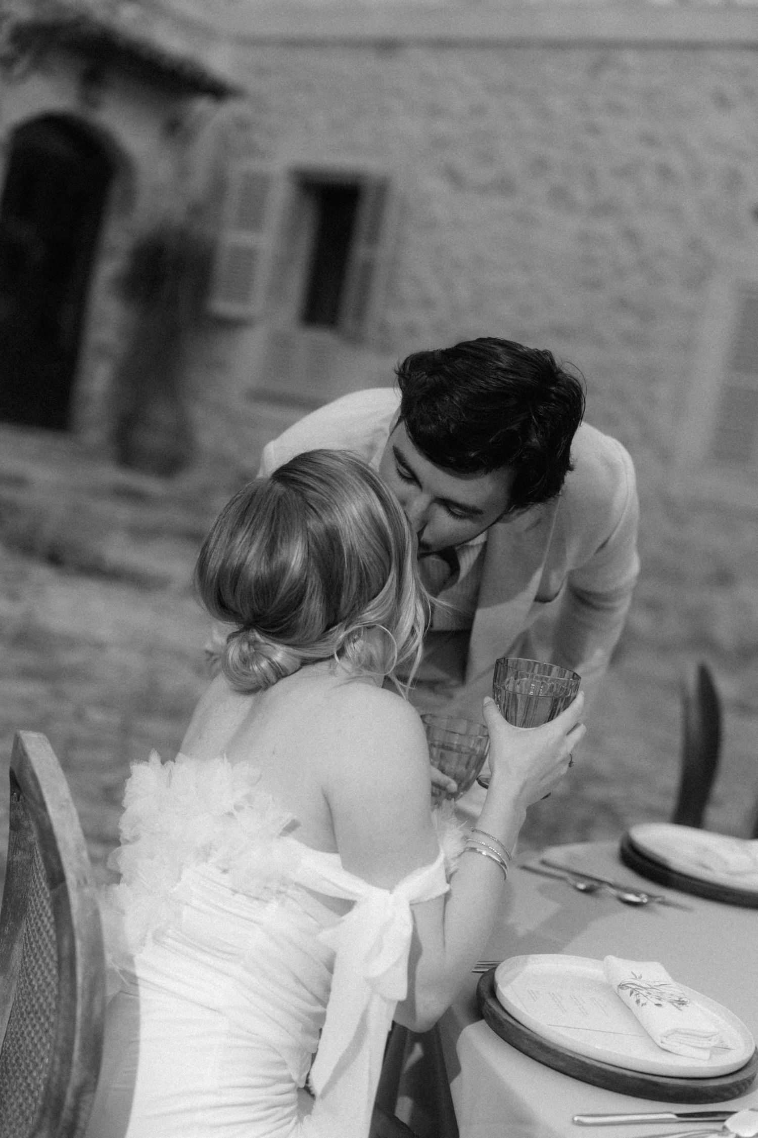 Black and white photo of groom leaning toward seated bride at outdoor wedding reception table.