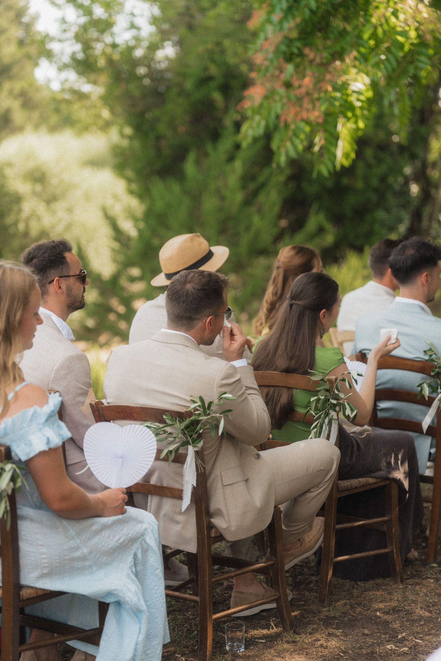 Wedding guests seated outdoors in garden ceremony, one wearing straw hat, holding heart-shaped fans on sunny day.