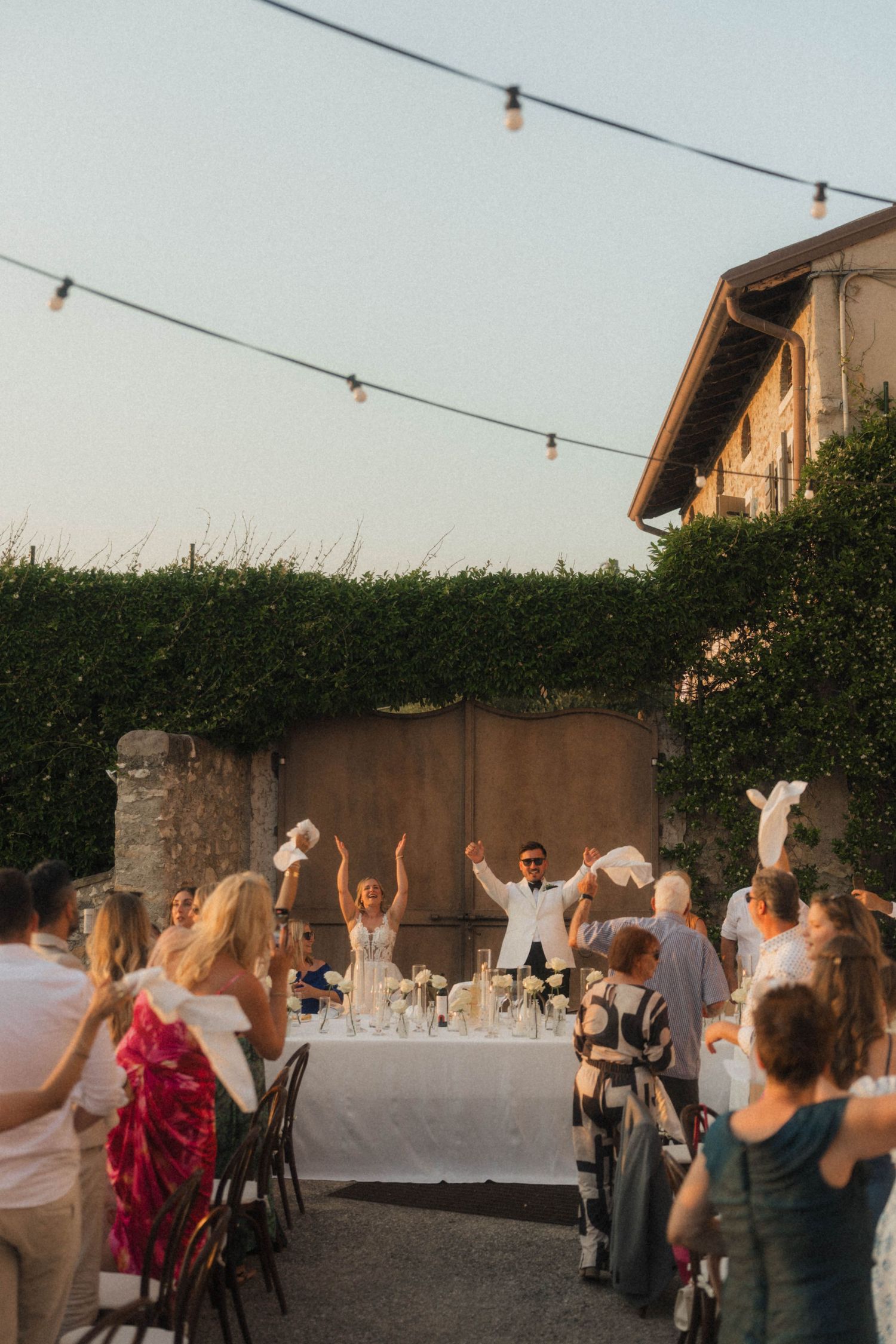 Wedding guests celebrating outdoors under string lights at rustic Italian venue at golden hour.