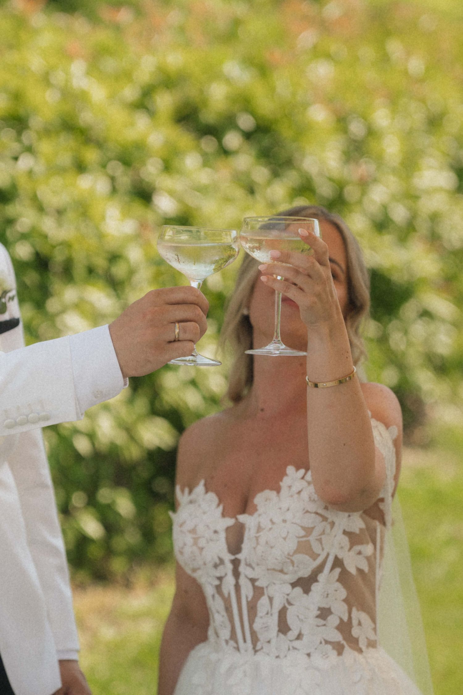 Bride and groom toasting with martini glasses at outdoor wedding ceremony surrounded by lush greenery.