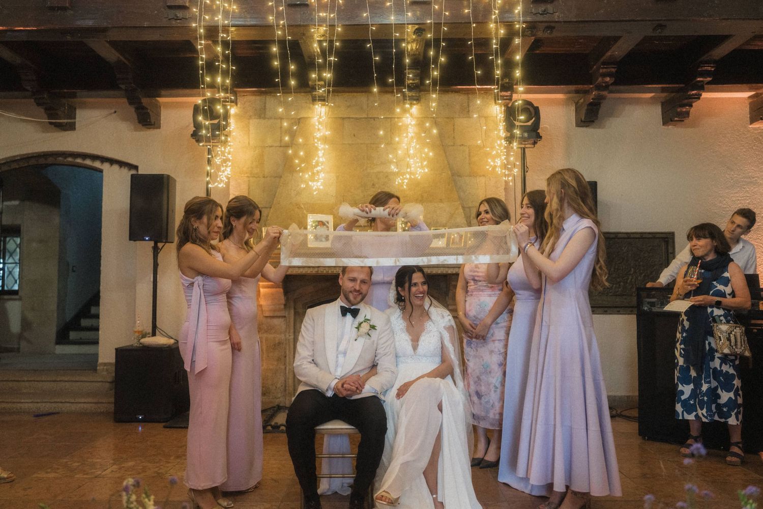 Bridesmaids in lavender dresses holding veil over bride and groom at romantic candlelit reception.