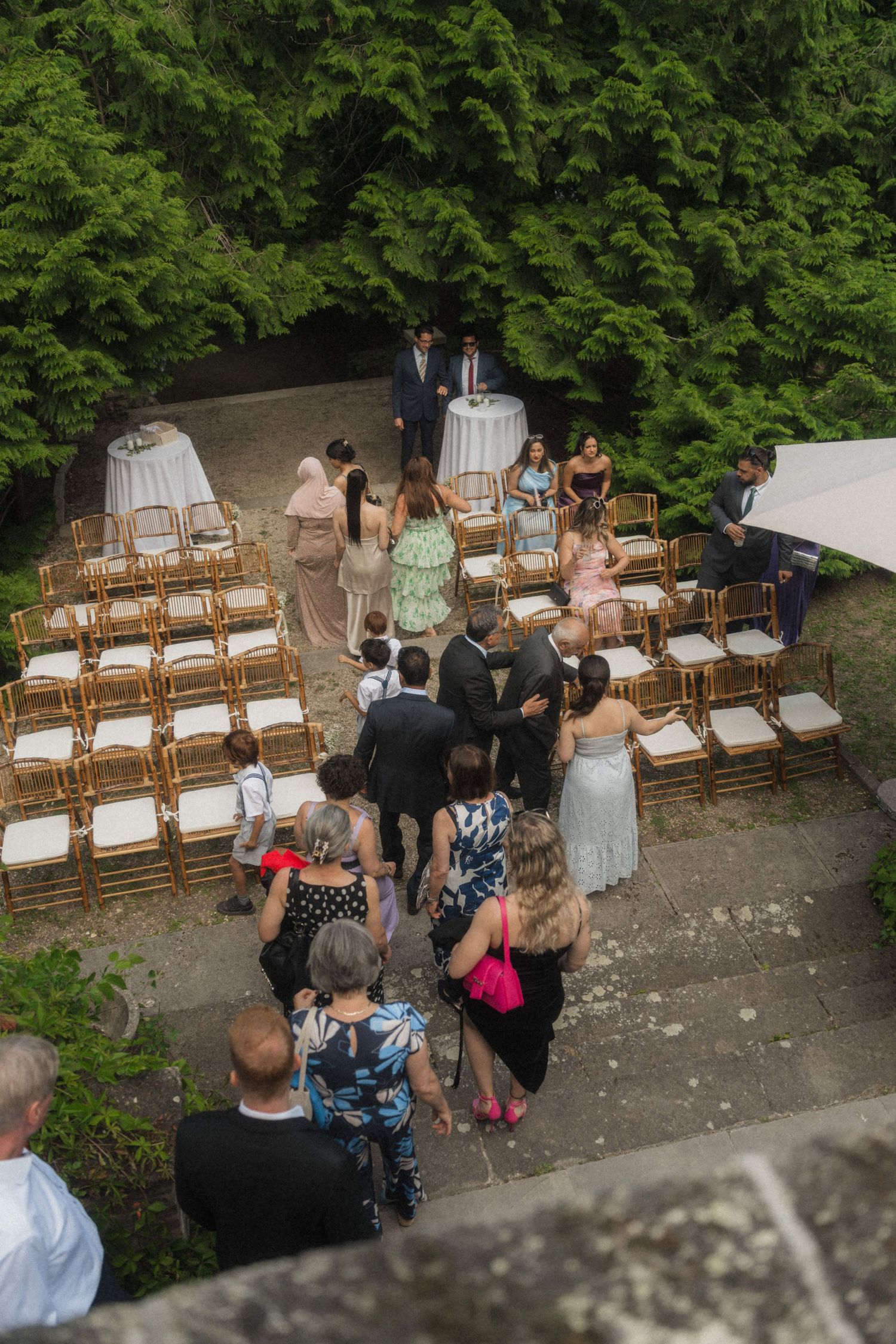Aerial view of wedding guests arriving at outdoor ceremony with rows of gold chairs surrounded by trees.