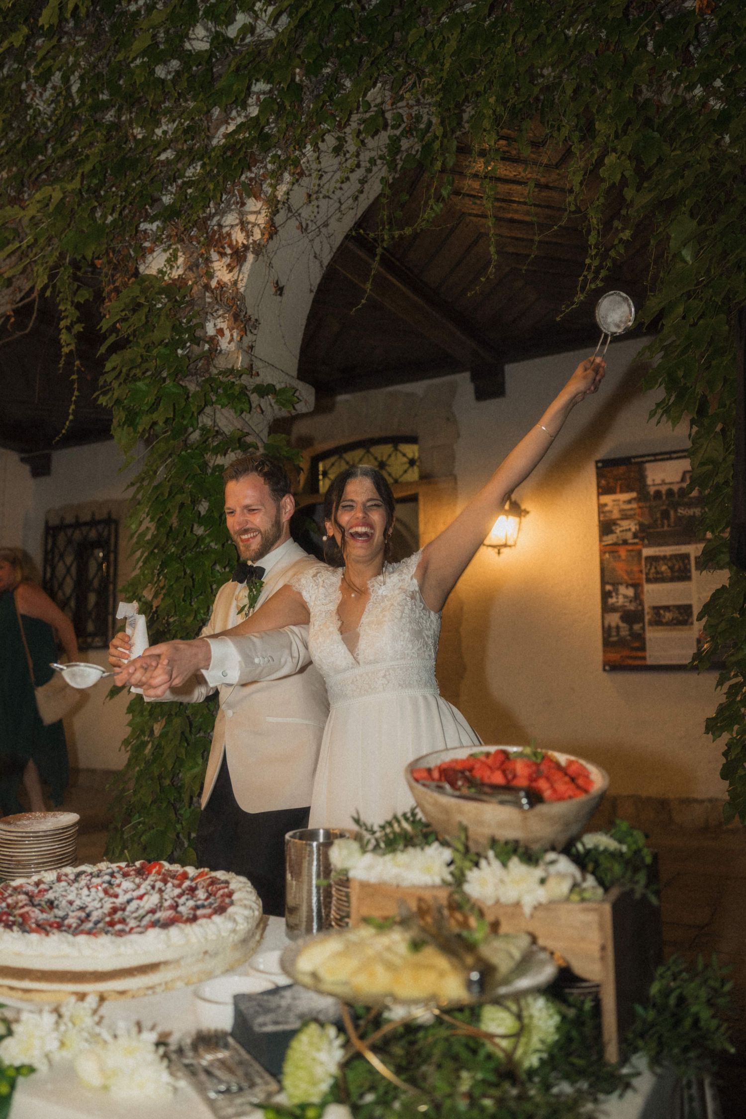 Joyful bride and groom cutting wedding cake at outdoor dessert table celebration.