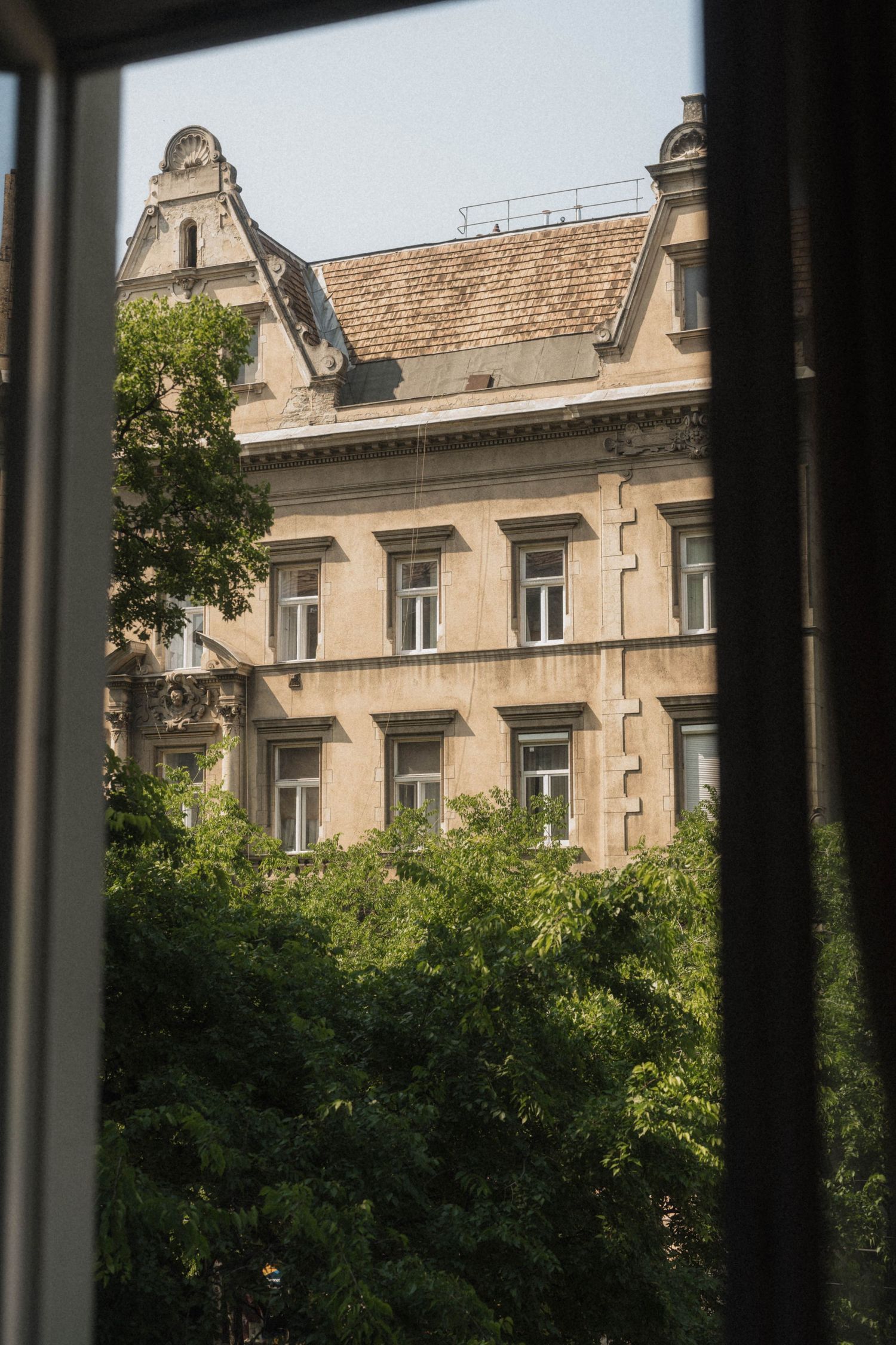 View through an open window of a historic European building facade with ornate gabled roof surrounded by lush green trees.