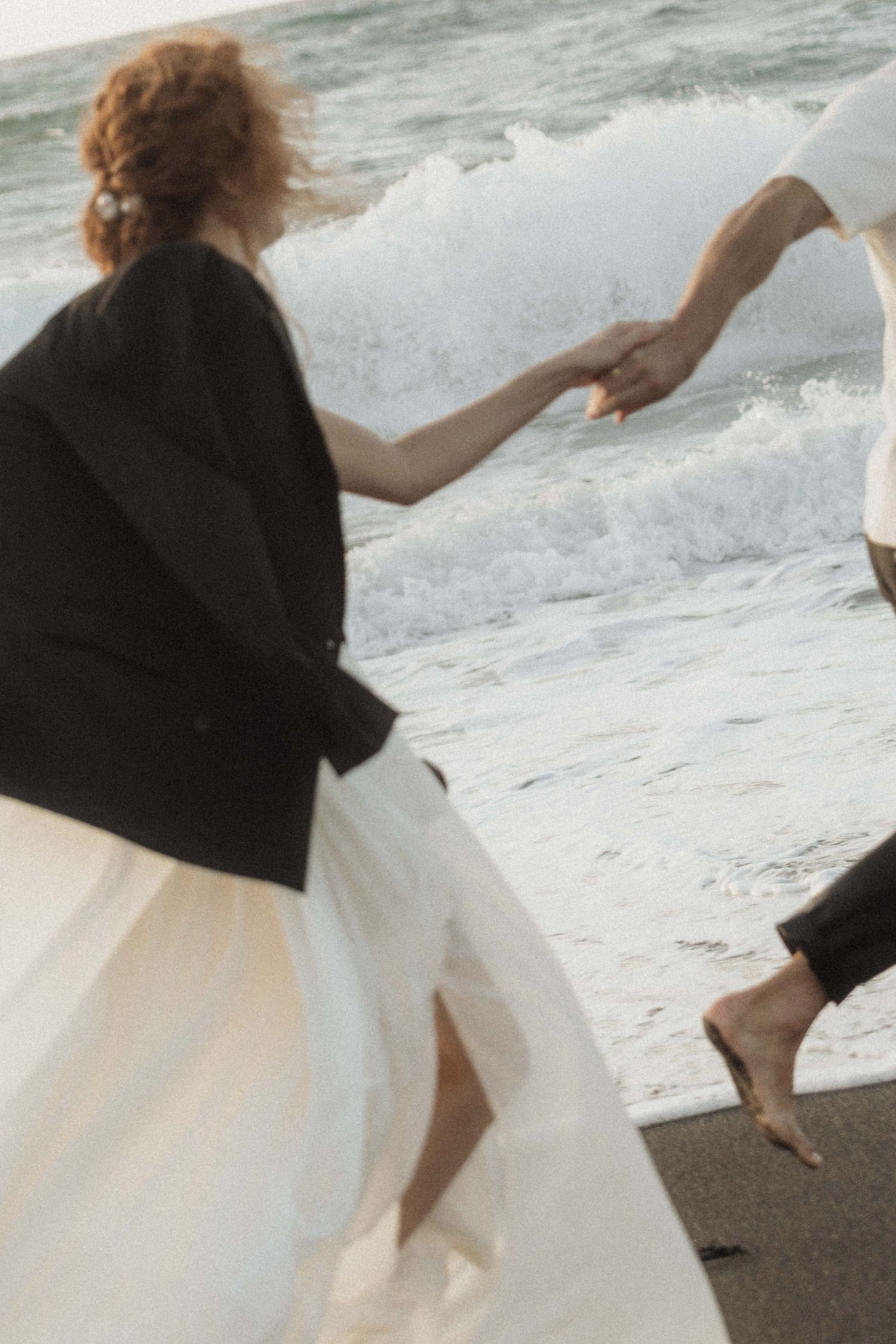 Bride in white skirt and black jacket holds groom's hand near crashing ocean waves on beach.