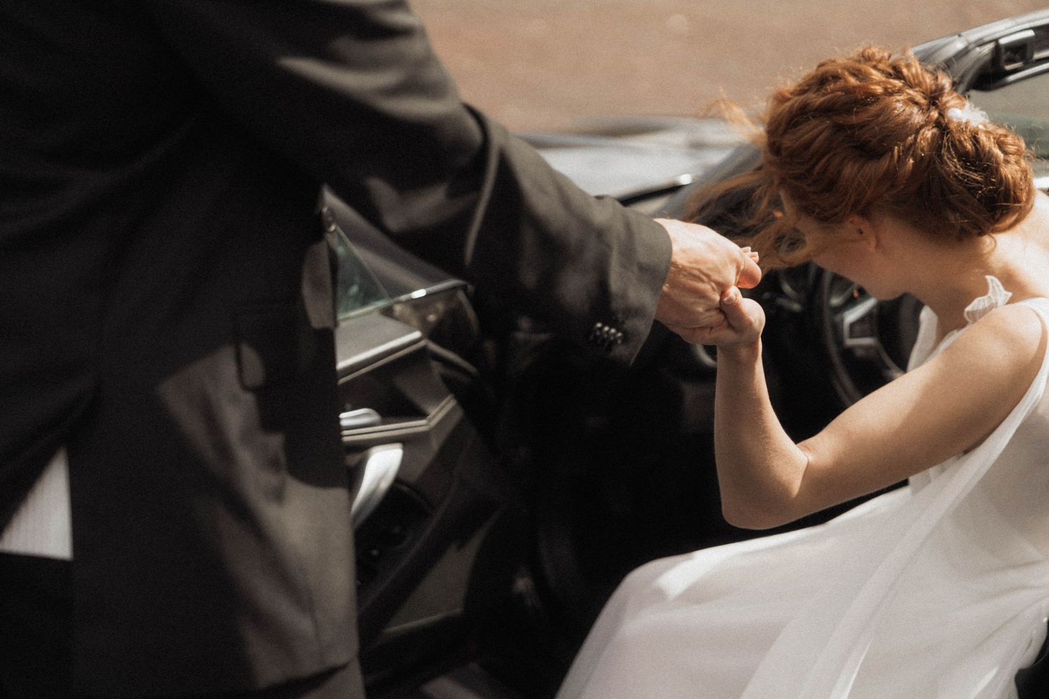 Bride in white dress being helped out of a black car by a groom in a suit, kissing his hand.