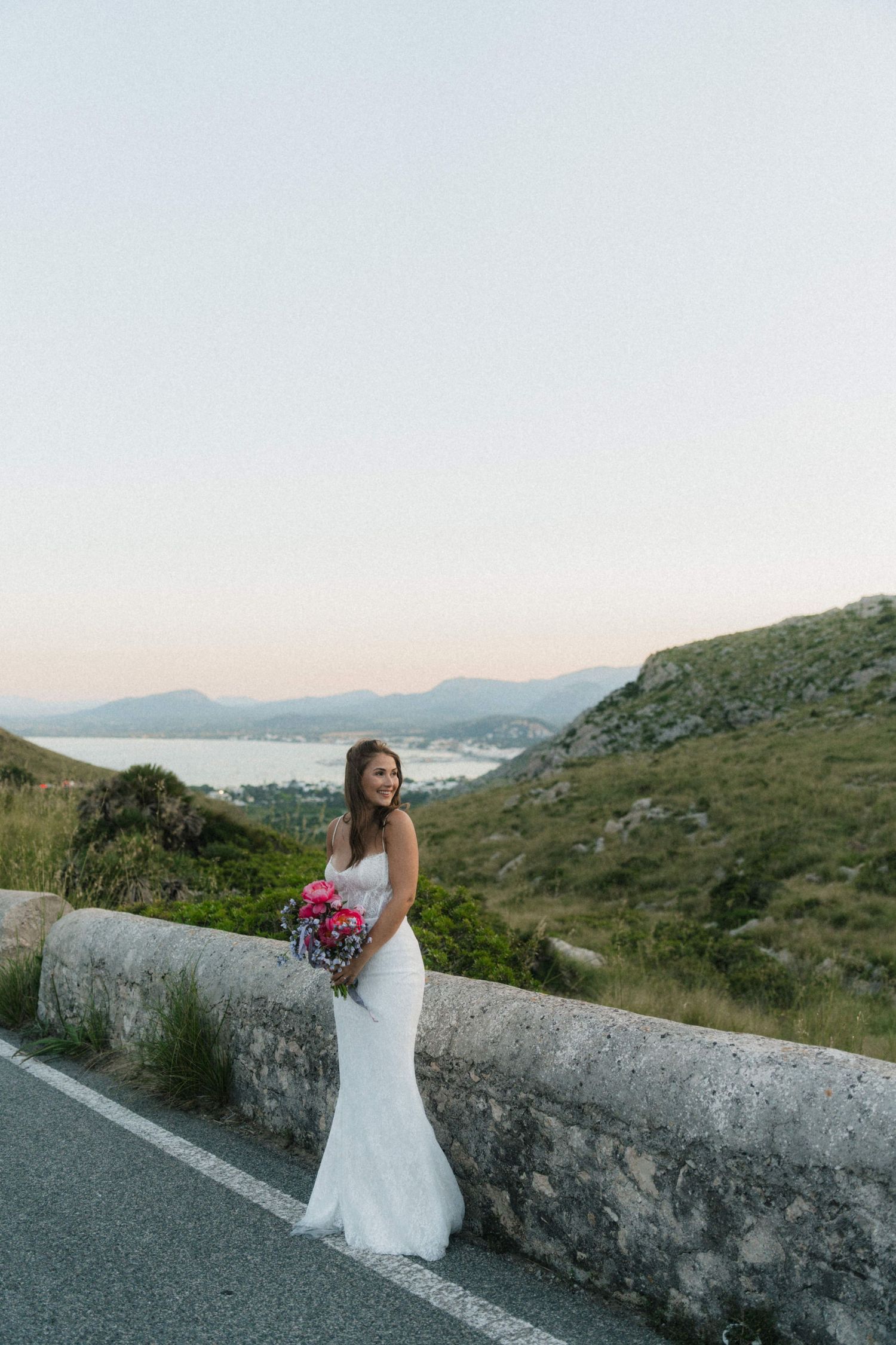 Bride in a fitted white lace gown posing by a stone road barrier with rolling hills and coast behind.