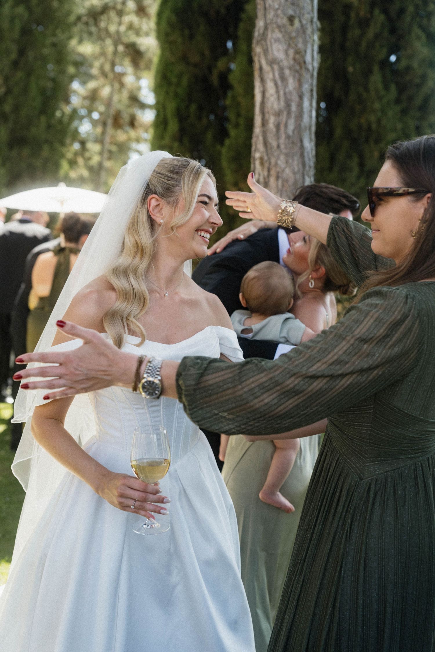 Smiling bride in white gown holding champagne receives warm greetings from wedding guests outdoors.