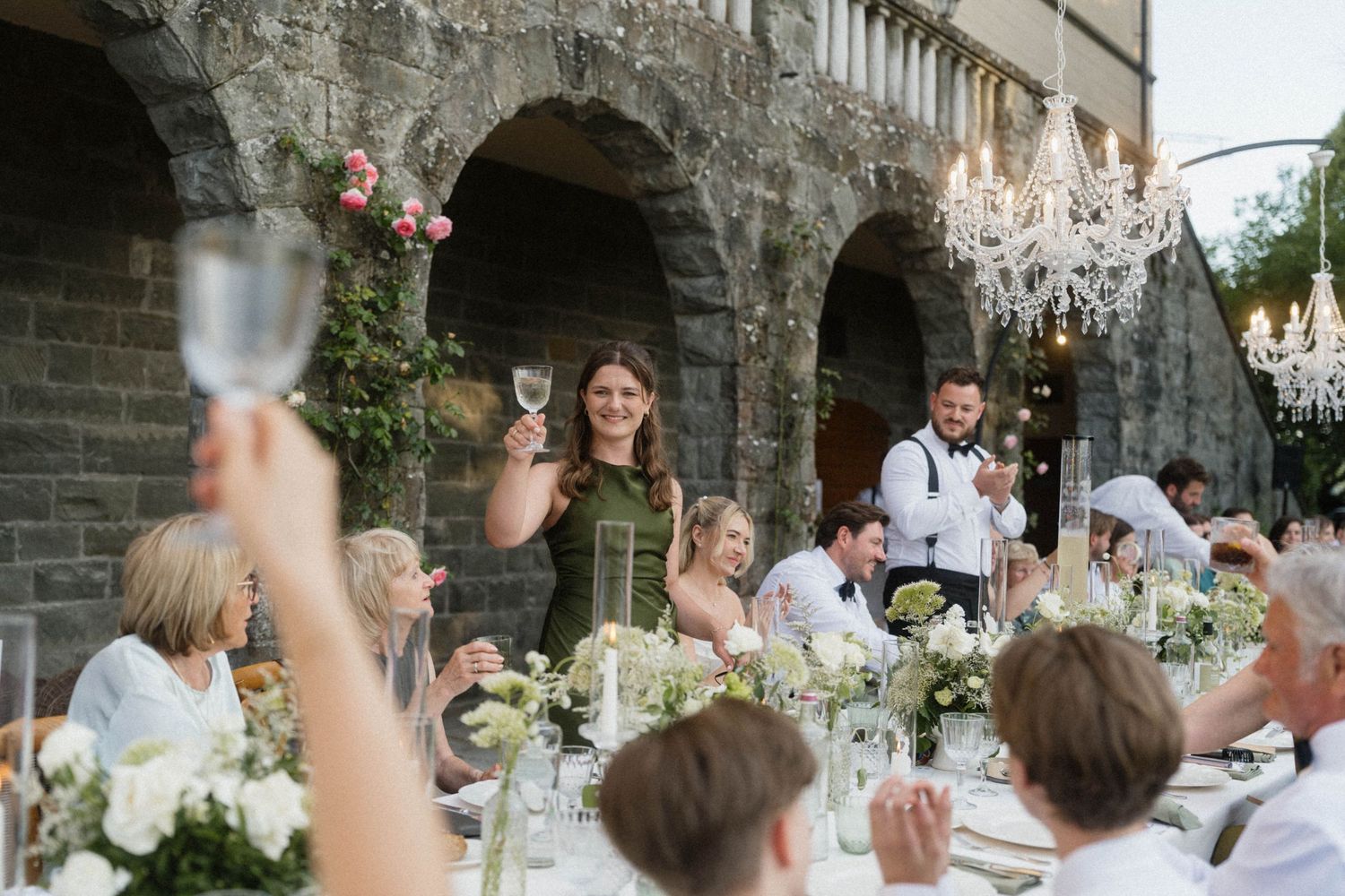Wedding guests raising glasses in a toast under crystal chandeliers at an elegant outdoor dinner.
