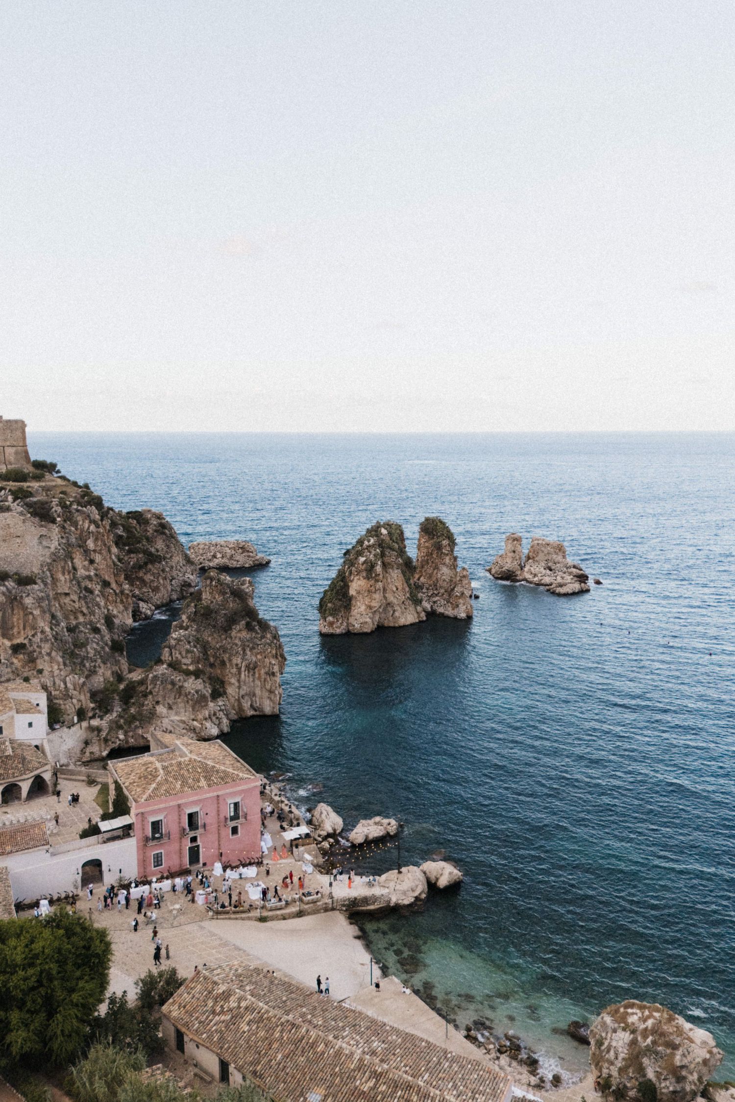Aerial view of a pink building on rocky cliffs beside turquoise Mediterranean sea with rock formations.