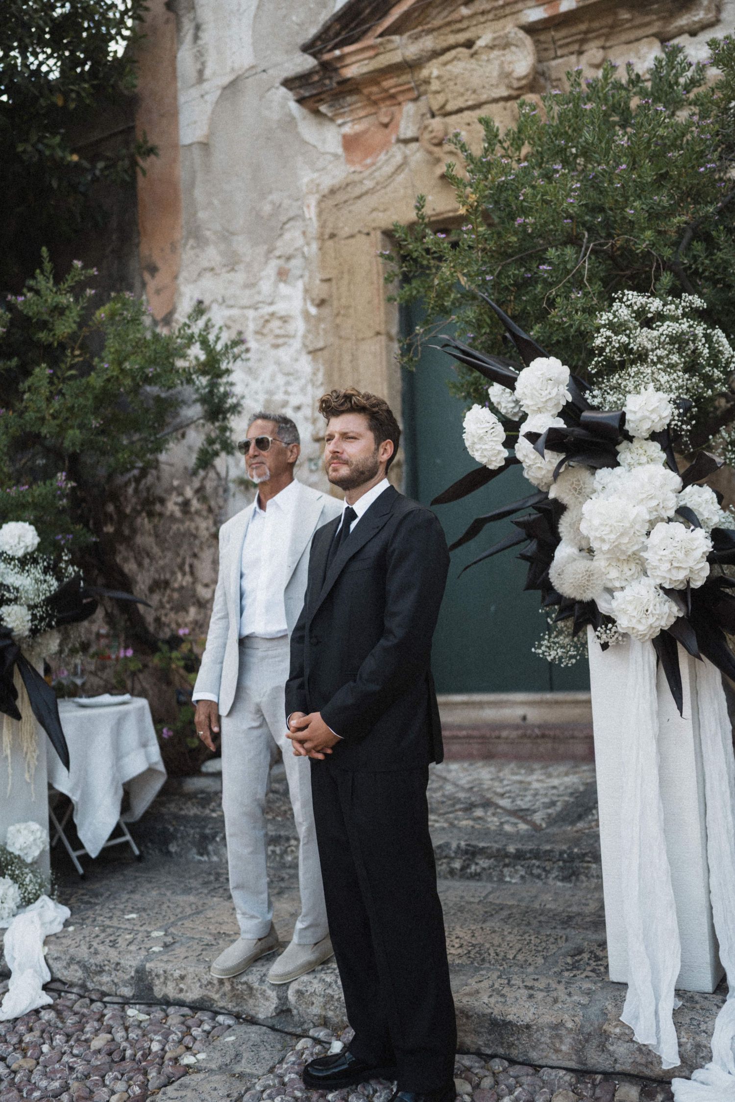 Groom in black suit standing at outdoor altar adorned with white floral arrangements.