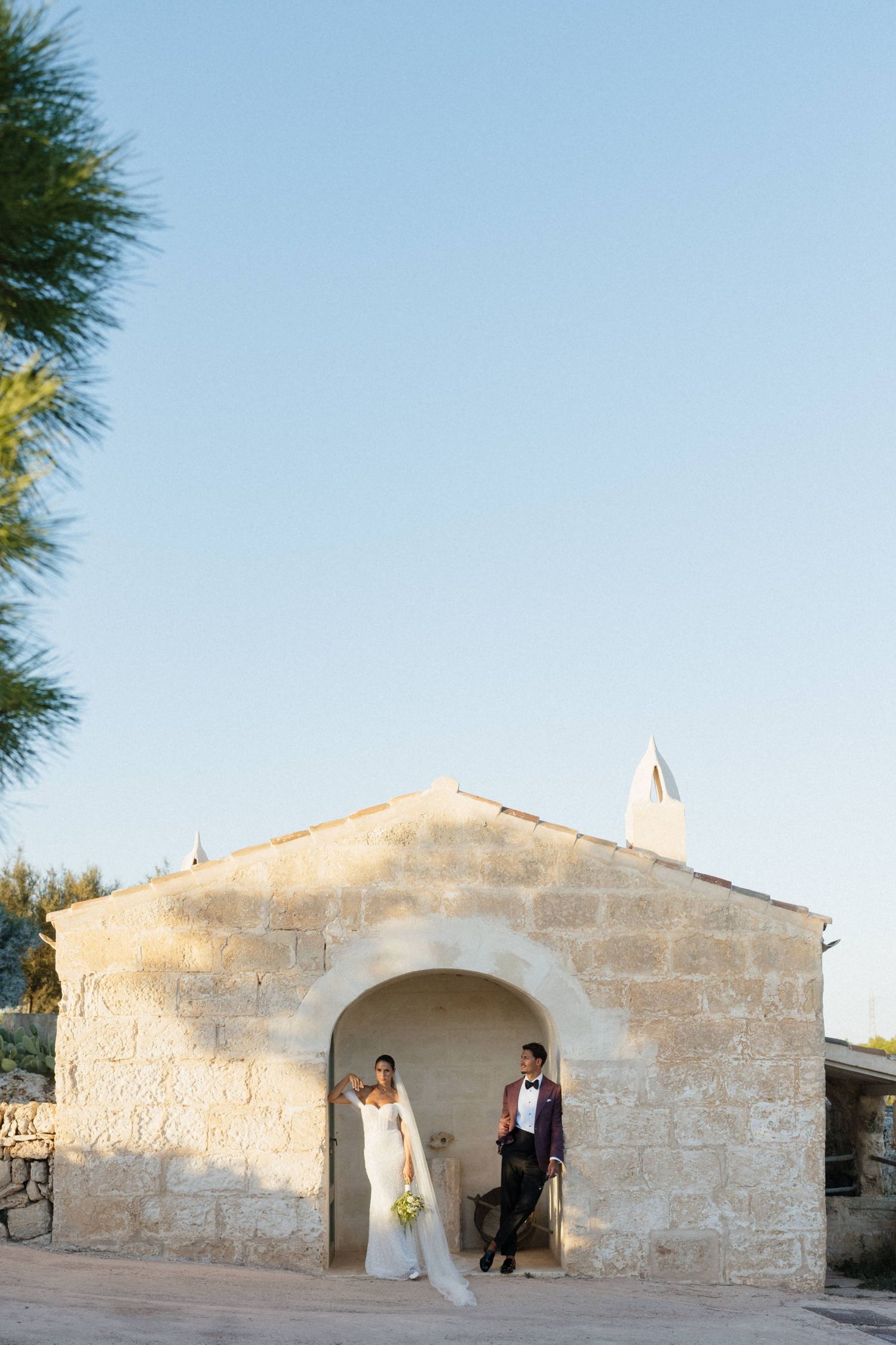 A bride and groom pose in the arched doorway of a rustic stone chapel under a clear blue sky at their wedding.