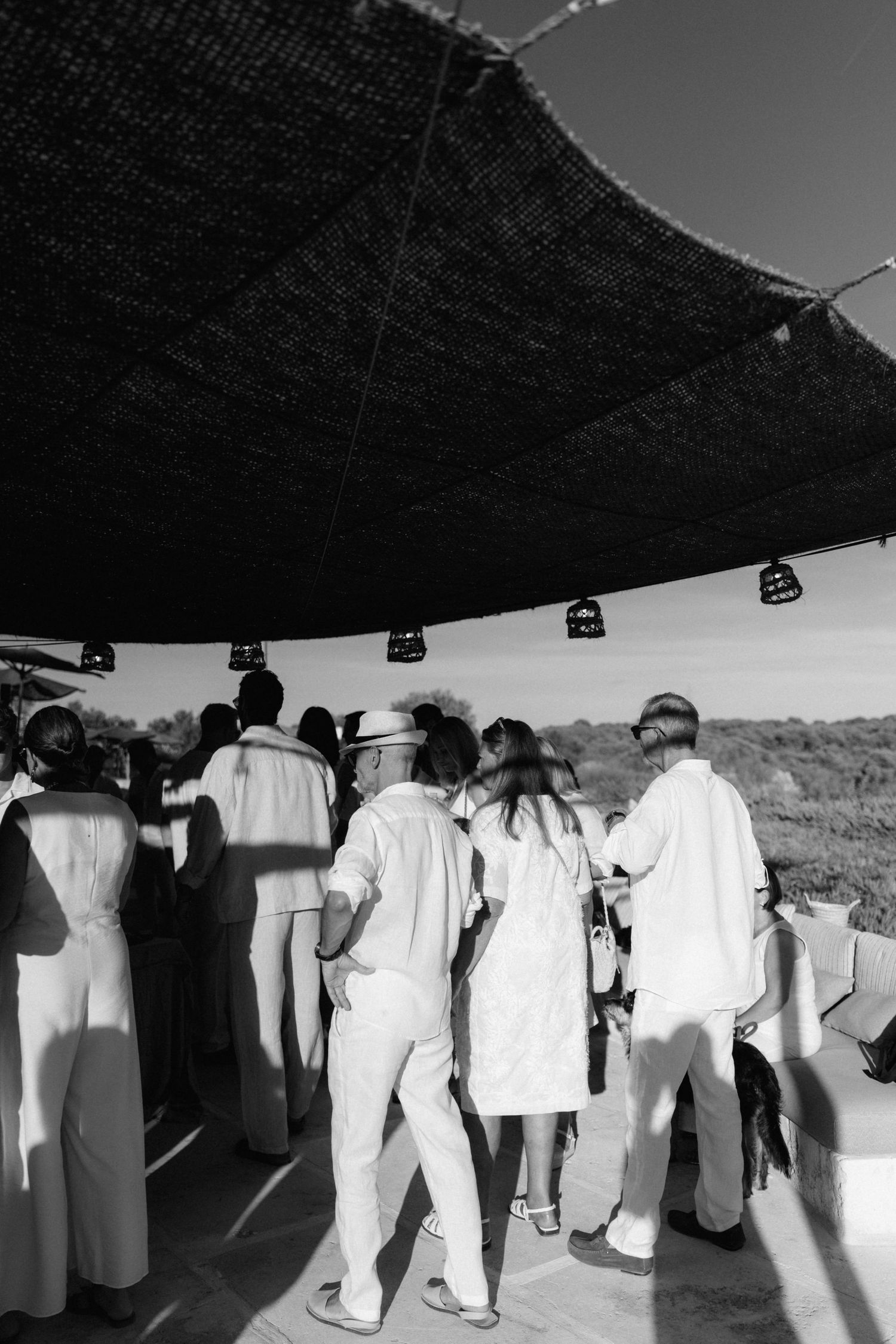 Guests in white linen outfits gather under a large woven shade canopy with hanging lanterns at an outdoor event in black and white.