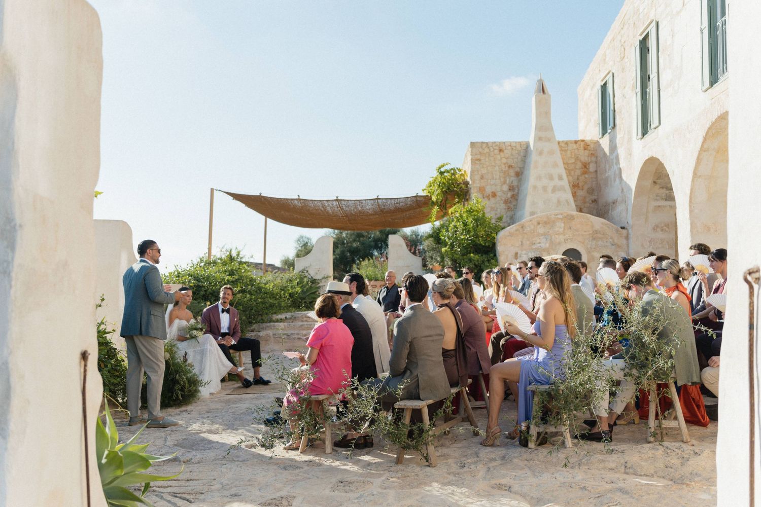 Guests sit on wooden benches under a woven canopy at an outdoor stone-courtyard wedding ceremony with a couple standing ahead.