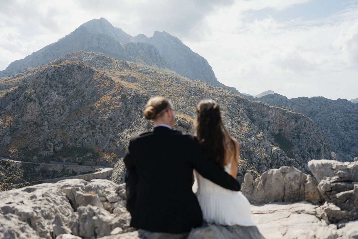 Couple in dark jacket and white dress sit on rocky ledge embracing while overlooking dramatic mountain valley under a cloudy sky.