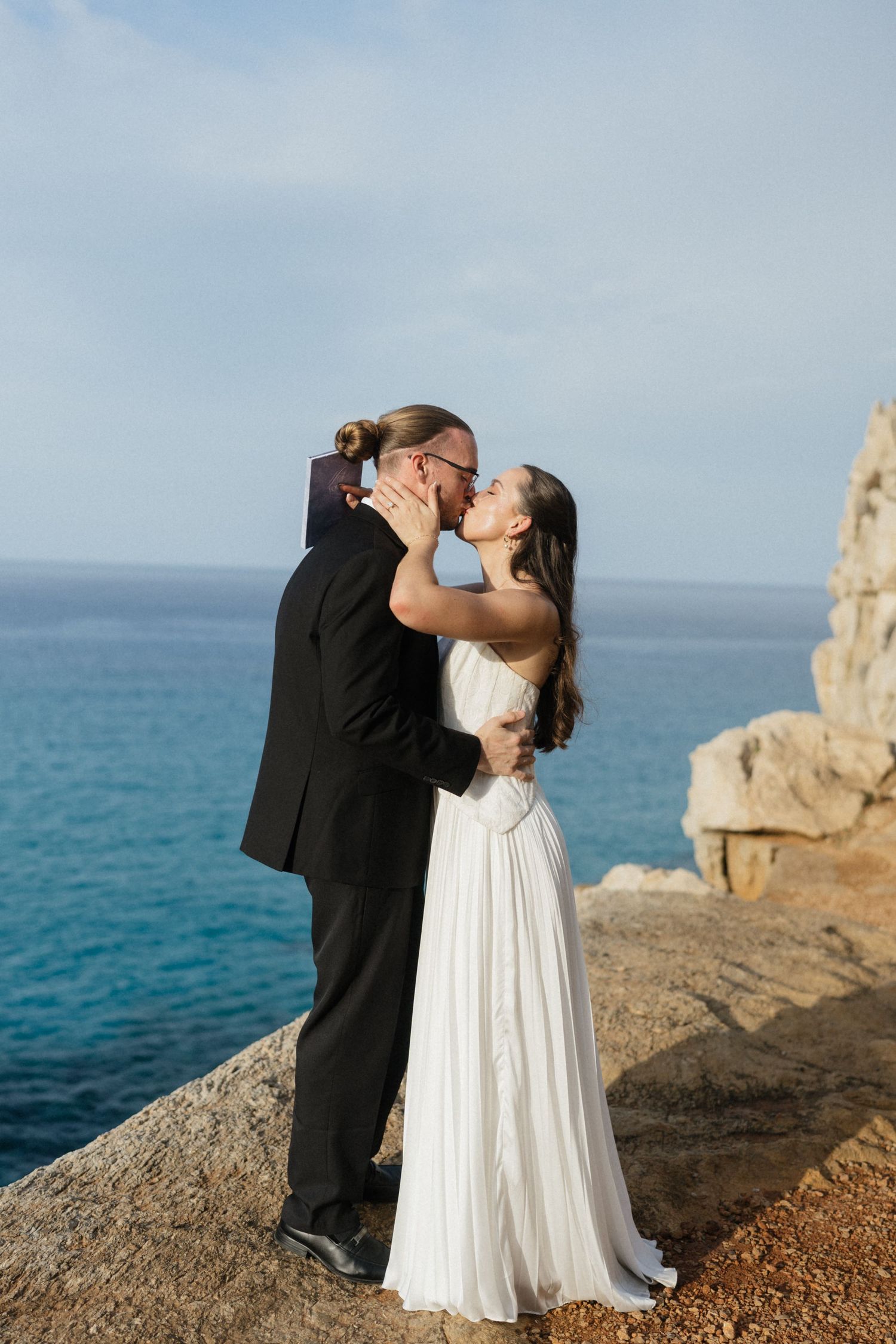 Bride and groom embrace in a romantic kiss on a sunlit coastal cliff with turquoise sea and rock formations behind them.