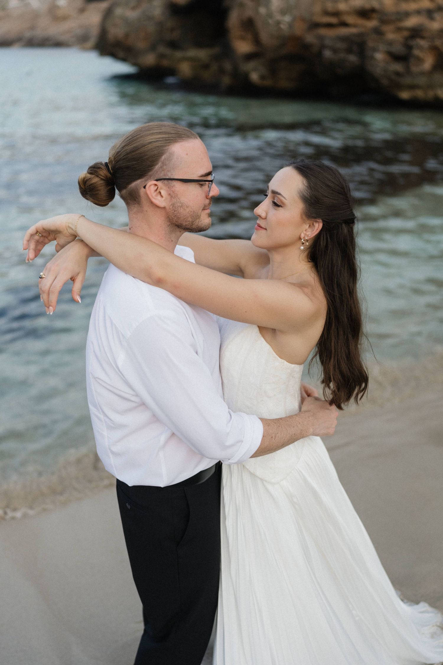 Bride in a white gown and groom in a white shirt embrace on a sandy beach beside calm turquoise water and rocky cliffs.