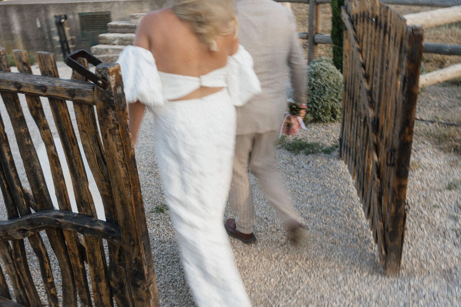 Bride in elegant white off-shoulder gown walking through rustic wooden gate with groom at wedding.