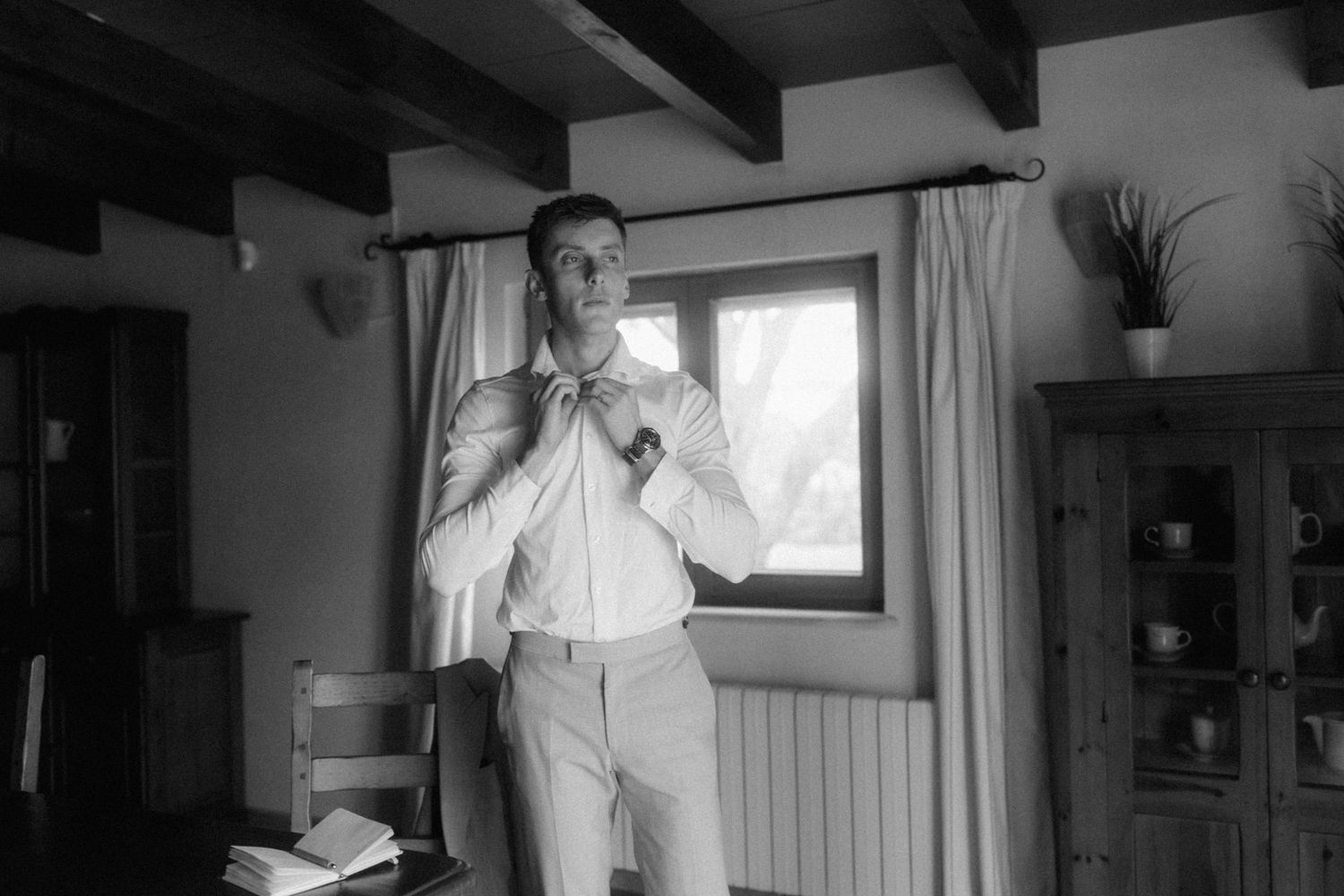 Black and white photo of a groom adjusting his tie while getting dressed in a rustic room before his wedding.