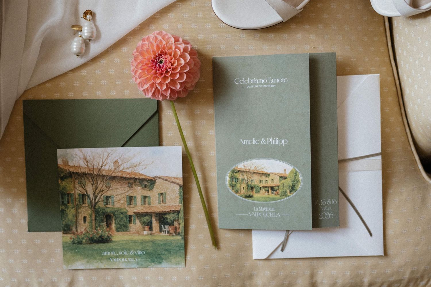 Elegant wedding stationery flat lay with sage green envelopes, watercolor venue illustration cards, and a pink dahlia flower.