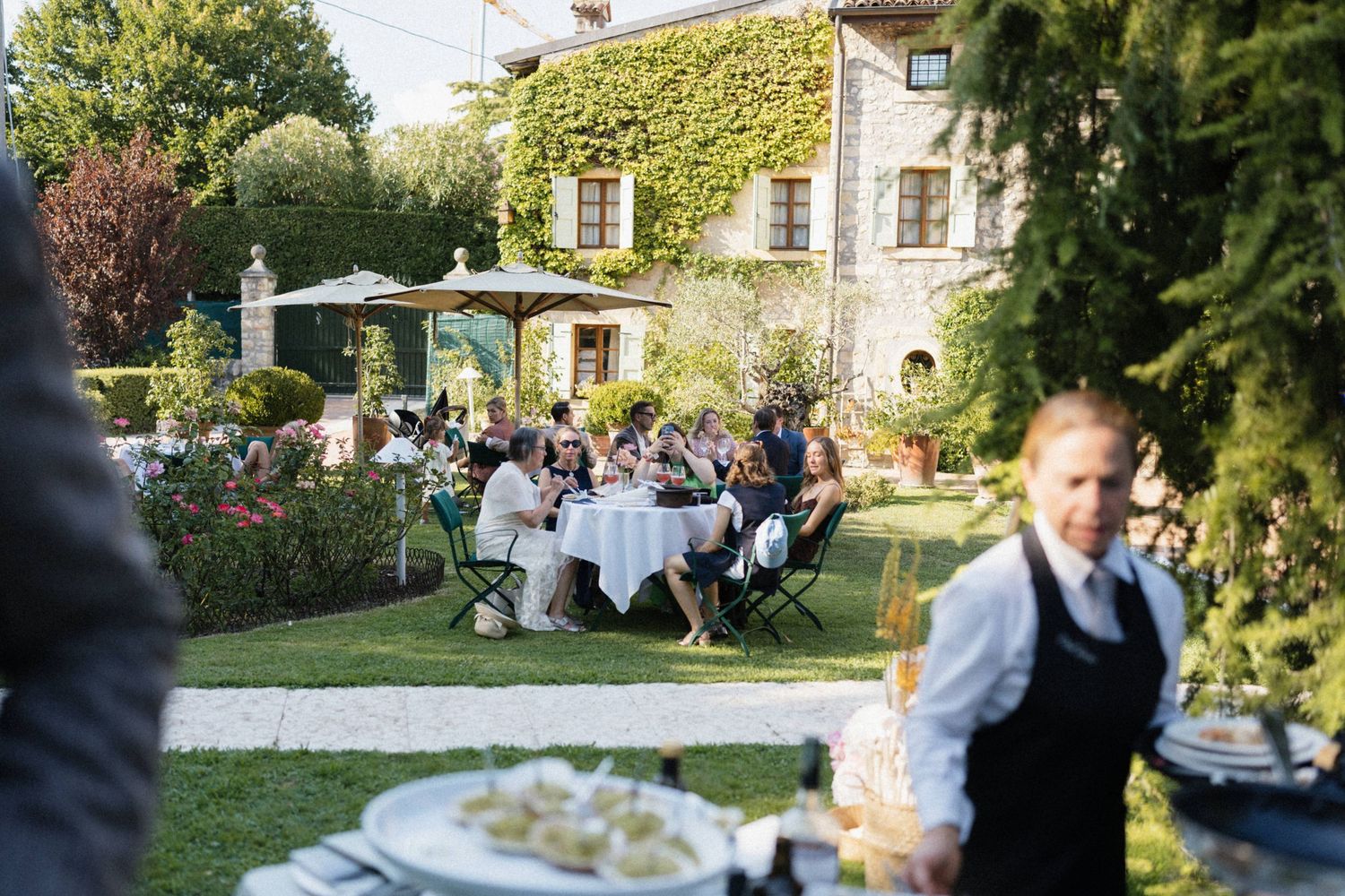 Elegant outdoor wedding reception in a lush Italian garden with guests dining at white-clothed tables beside a stone villa.