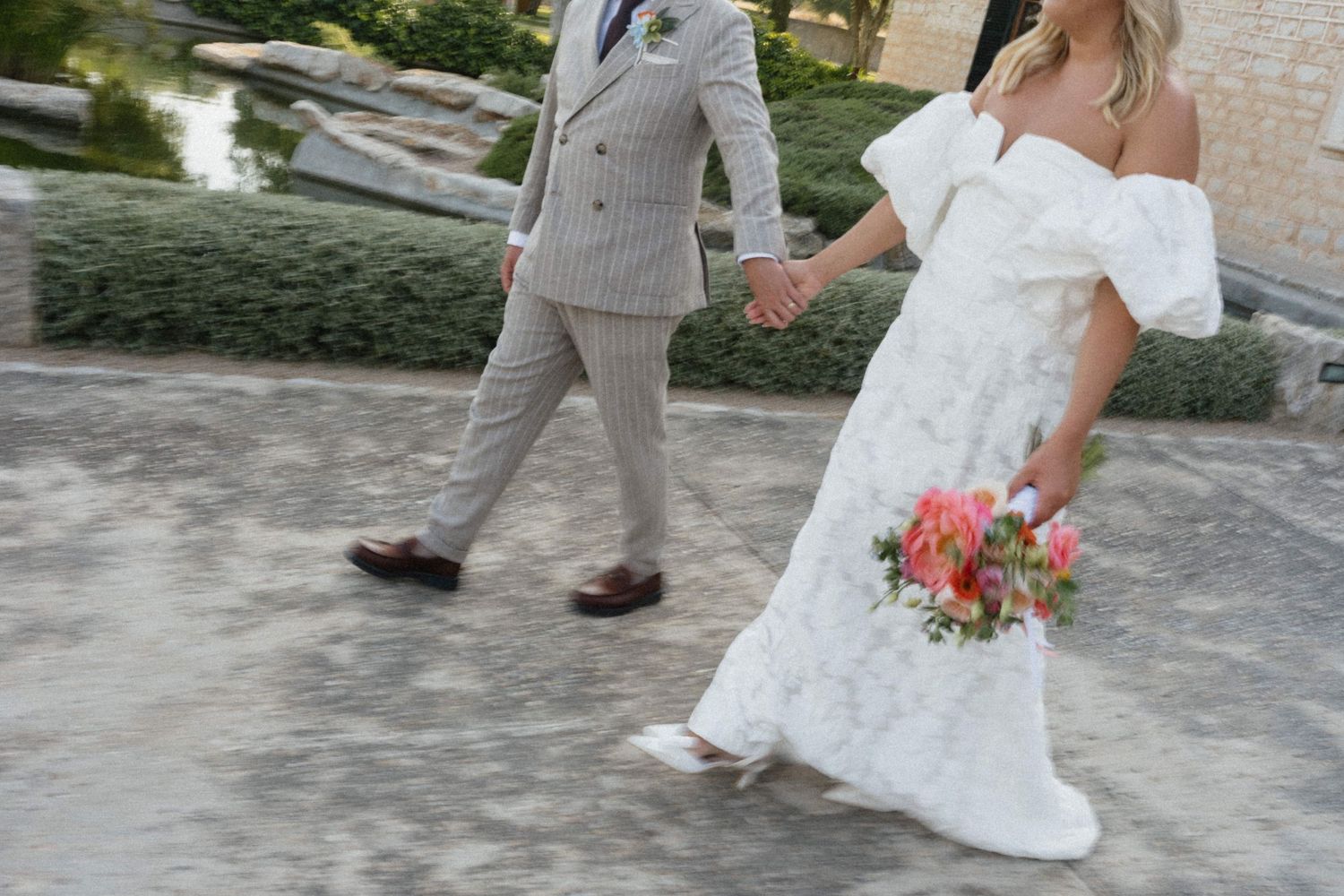 Bride in white off-shoulder gown holding pink bouquet walks hand-in-hand with groom in gray pinstripe suit outdoors.