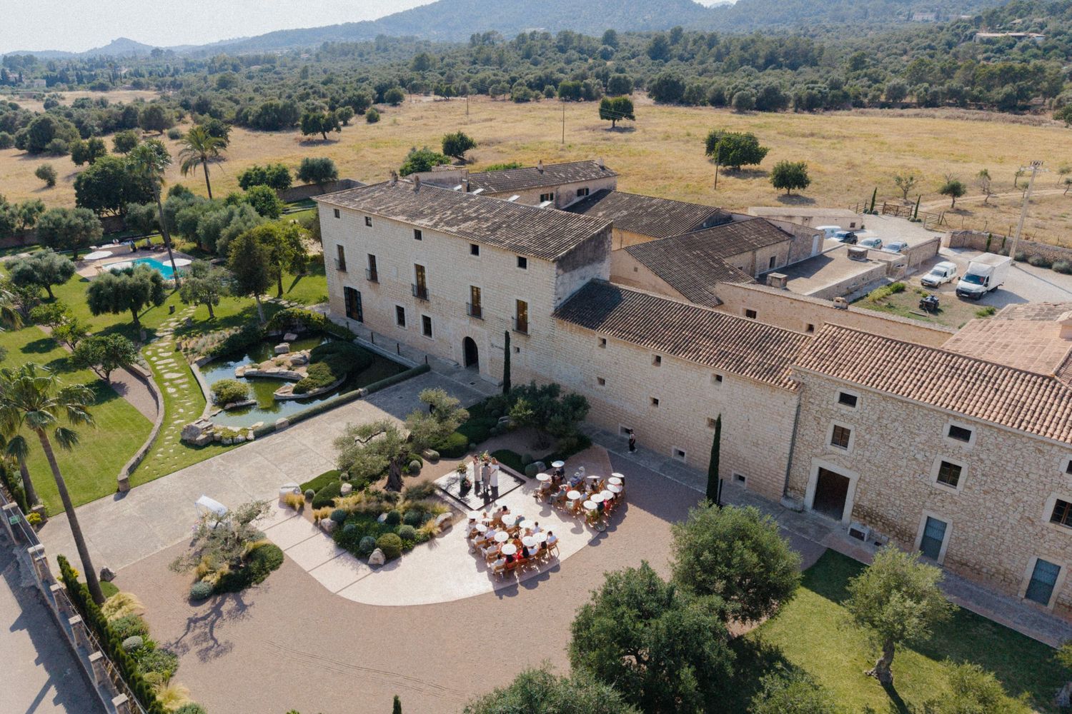 Aerial view of a historic stone manor house with terracotta roof, courtyard dining, pool, and lush gardens in a rural landscape.