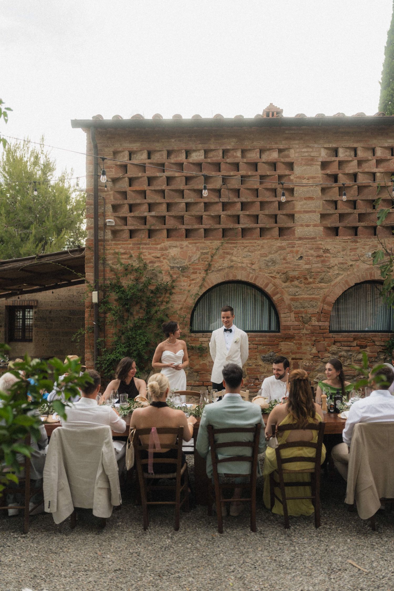 Bride and groom address guests at an elegant outdoor wedding reception beside a historic Italian brick villa.