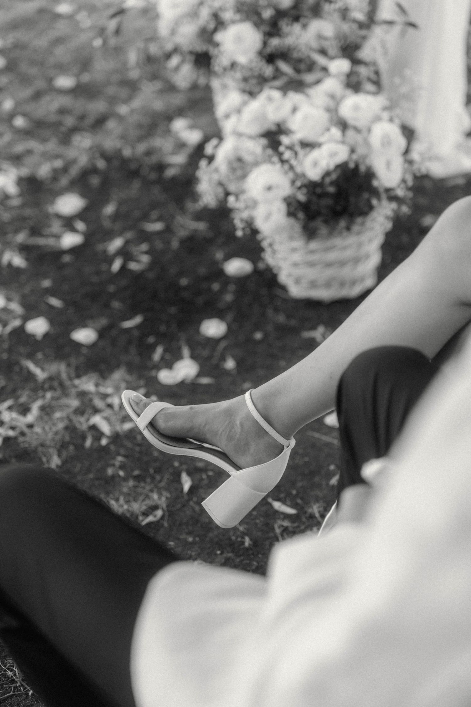 Black and white photo of bride's white heeled sandals with floral arrangement in background.