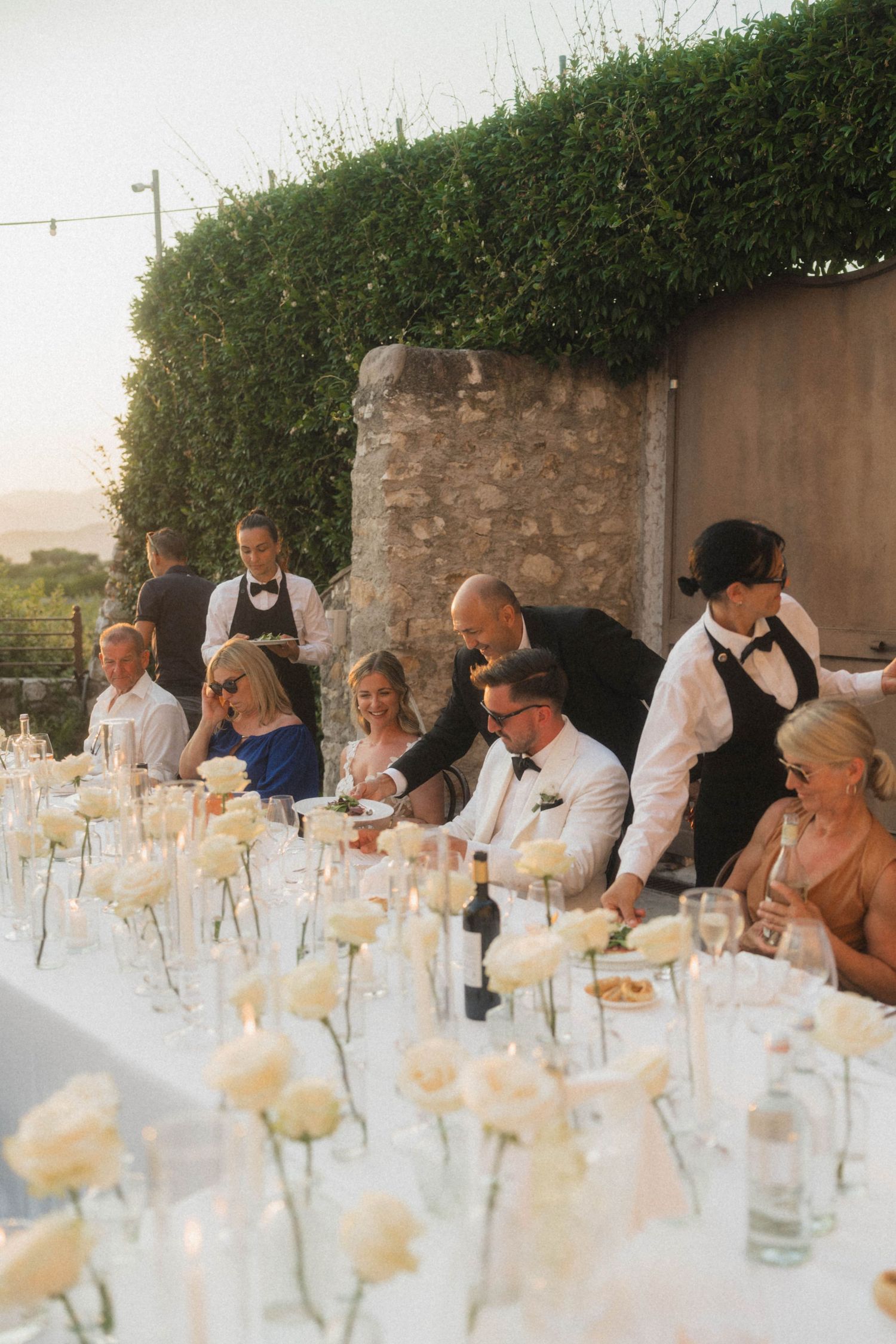 Wedding guests dining at elegant outdoor reception table with white roses and candles.