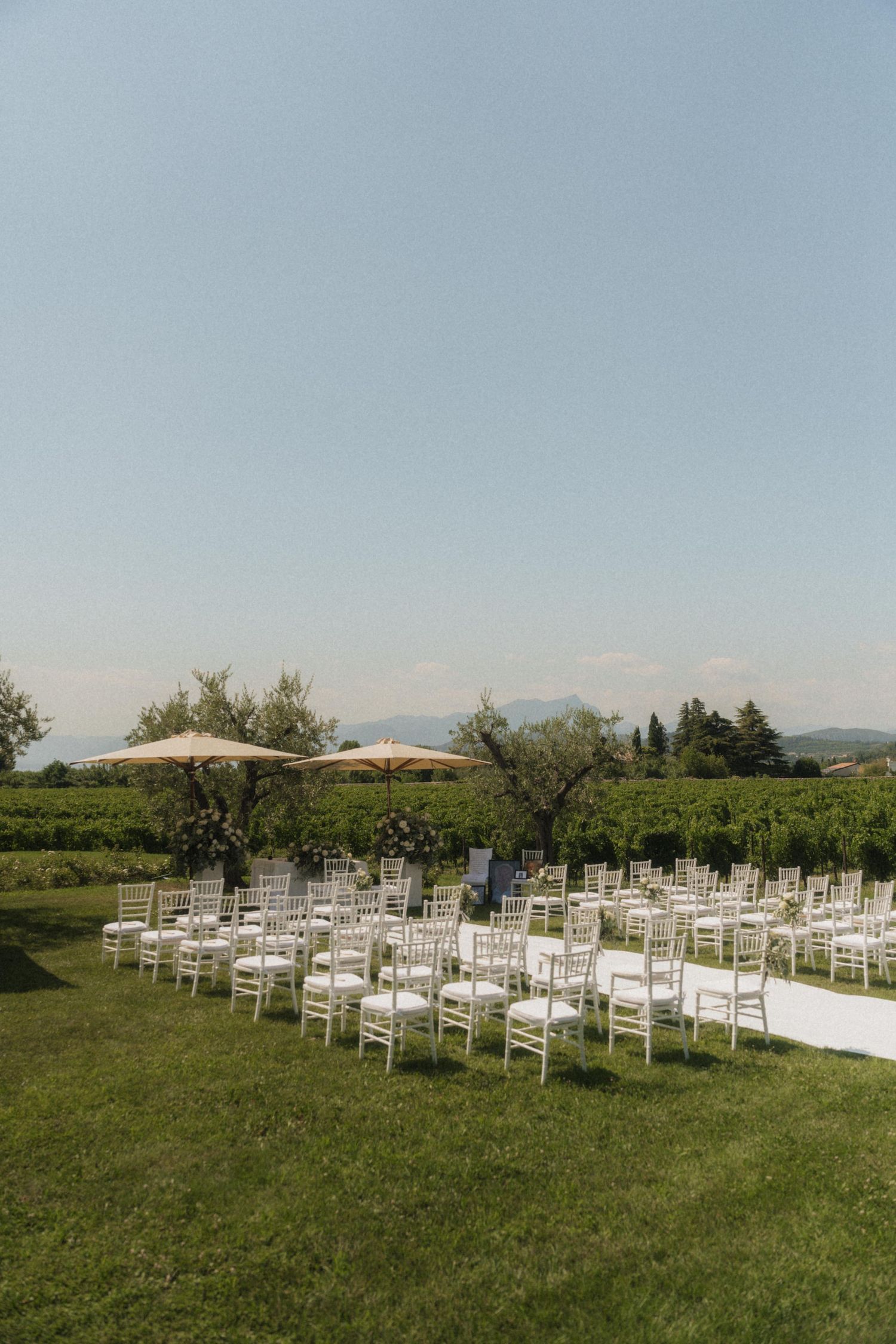 Elegant outdoor wedding ceremony setup with white chiavari chairs on green lawn, vineyard backdrop, and mountain views under clear blue sky.
