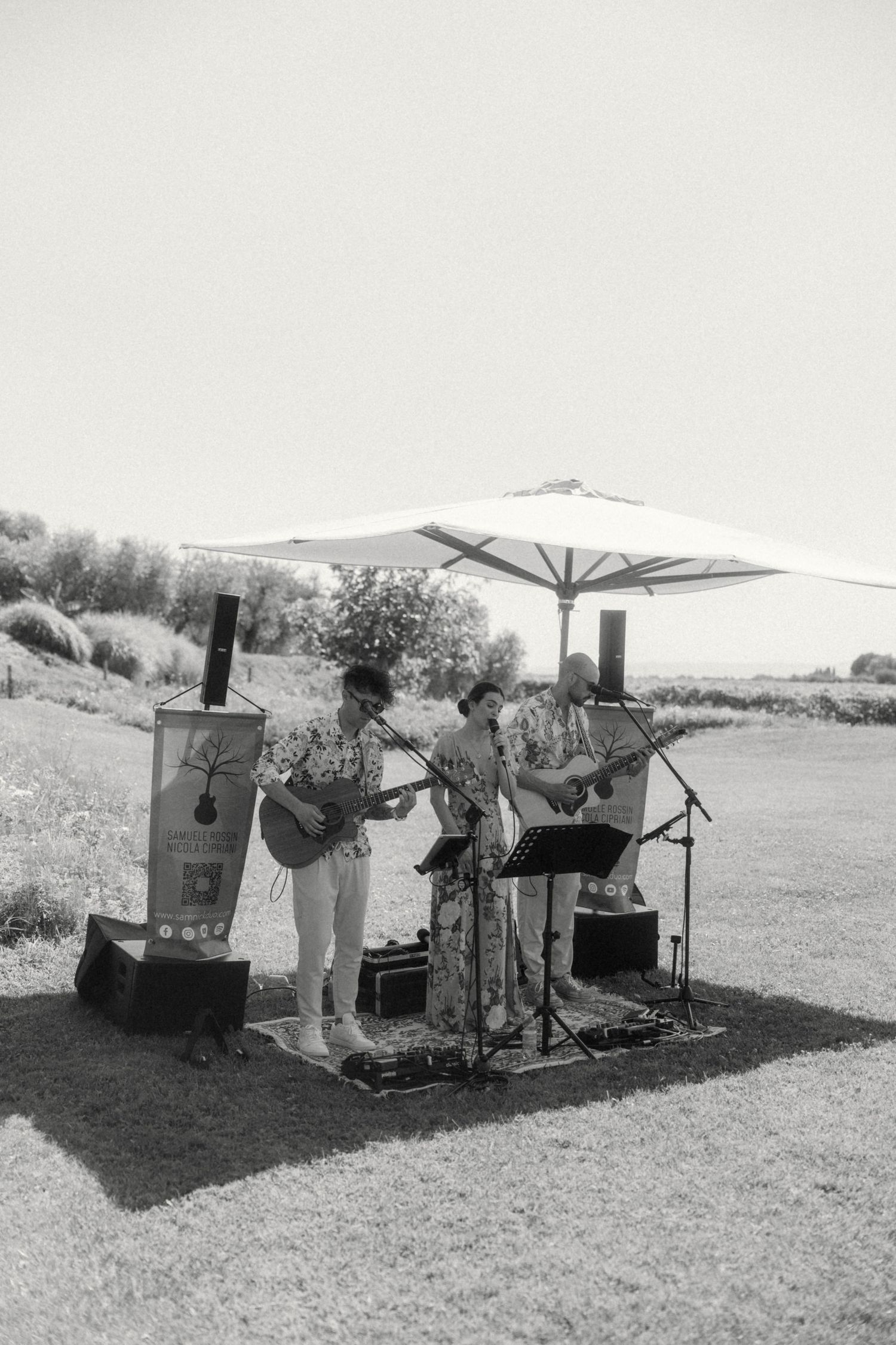 Black and white photo of a three-piece band performing outdoors under a white umbrella at a vineyard wedding.