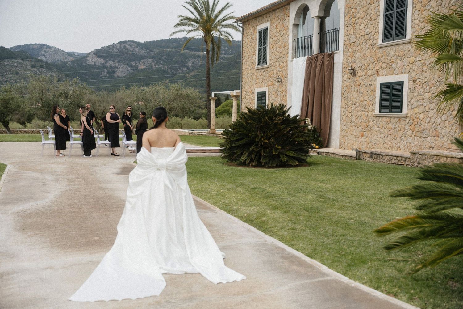 Bride in flowing white off-shoulder gown walking toward bridesmaids in black dresses at rustic estate.