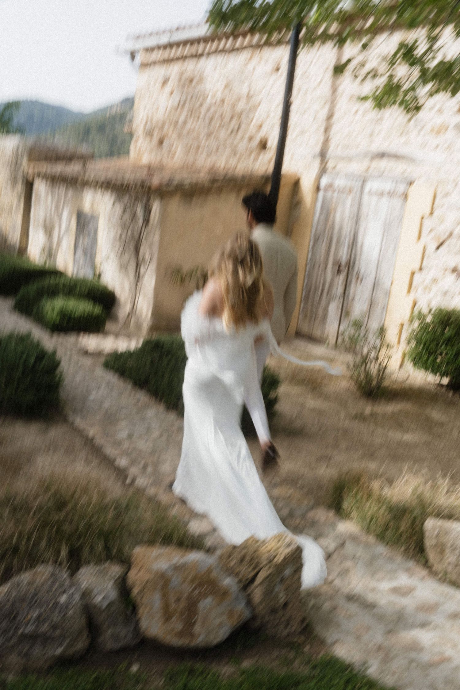 Bride in white gown and groom walking along stone path near rustic building in blurred motion photo.