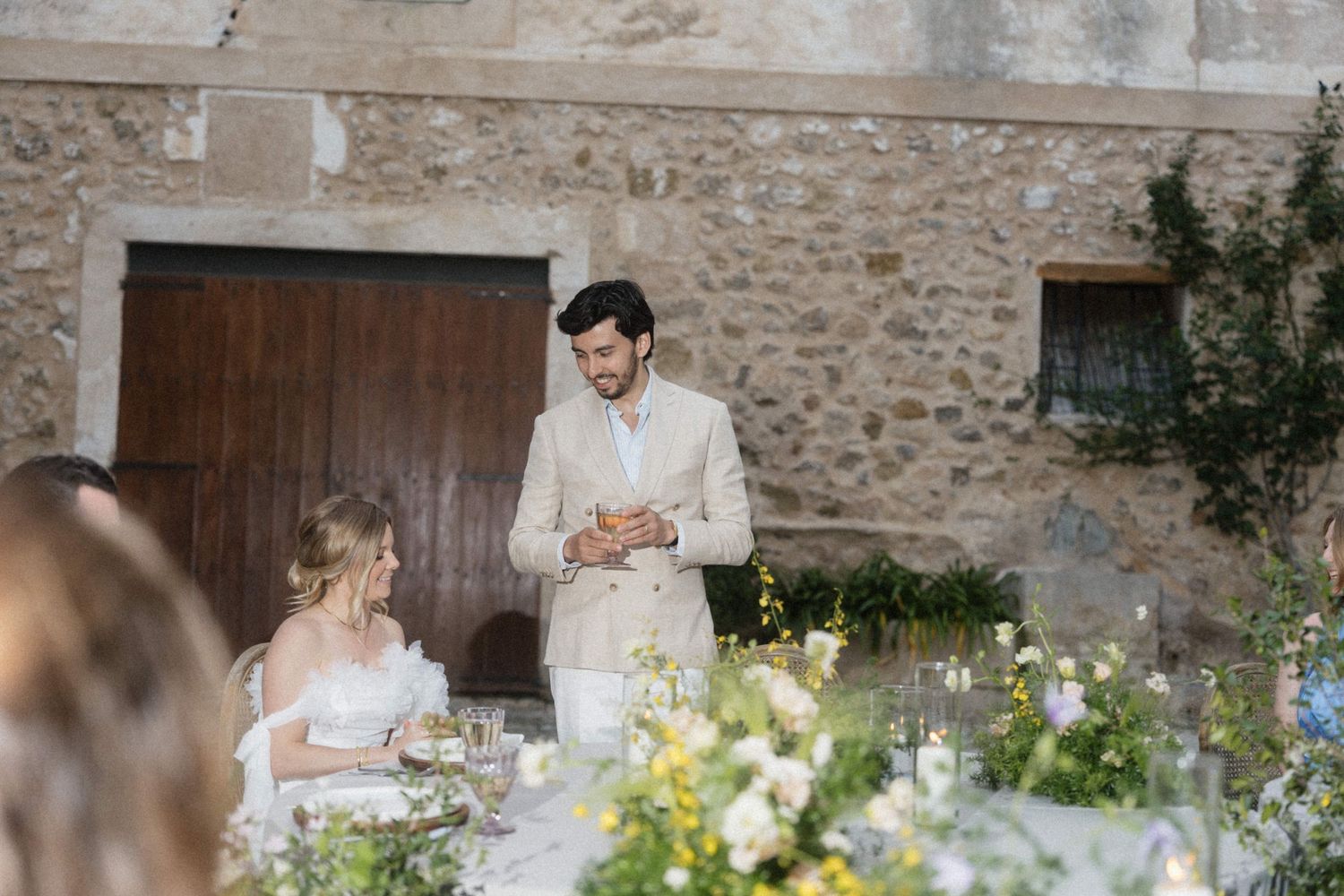 Groom in beige suit stands speaking to bride at floral-decorated outdoor wedding reception table.