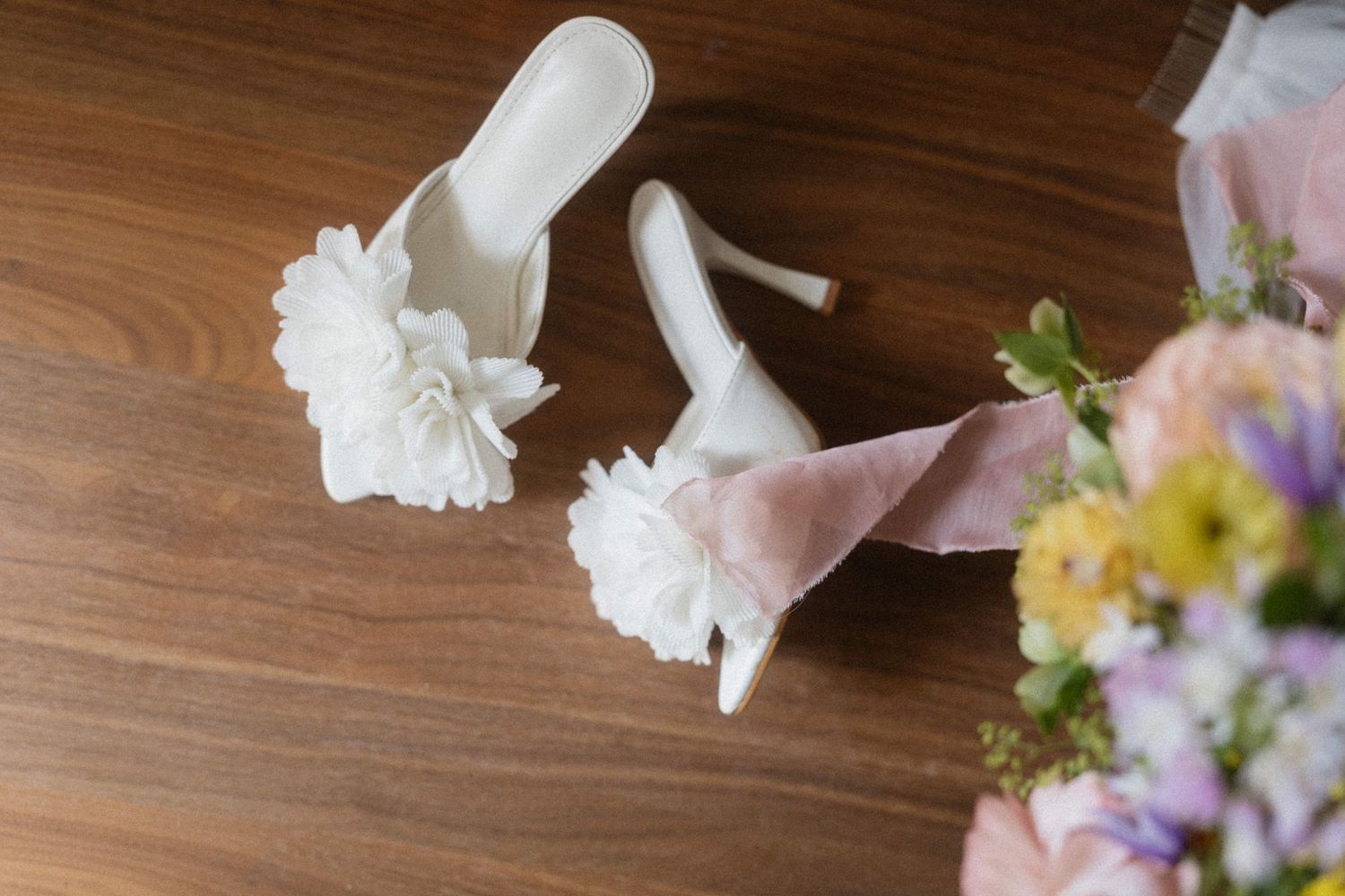 White bridal heeled mules with floral embellishments on wooden floor beside a pastel wedding bouquet.