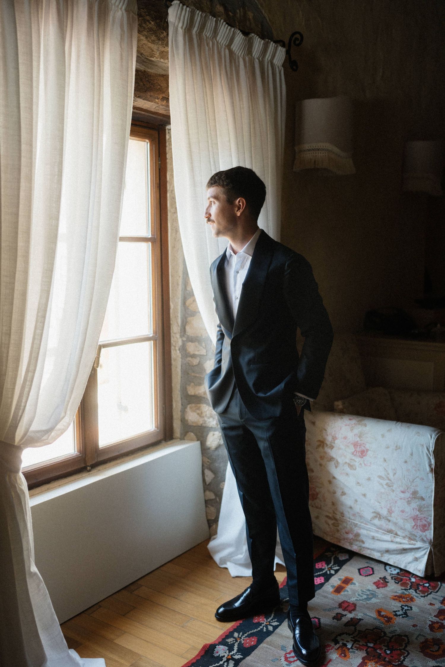 Elegant groom in dark suit standing by window with curtains looking outside on wedding day.