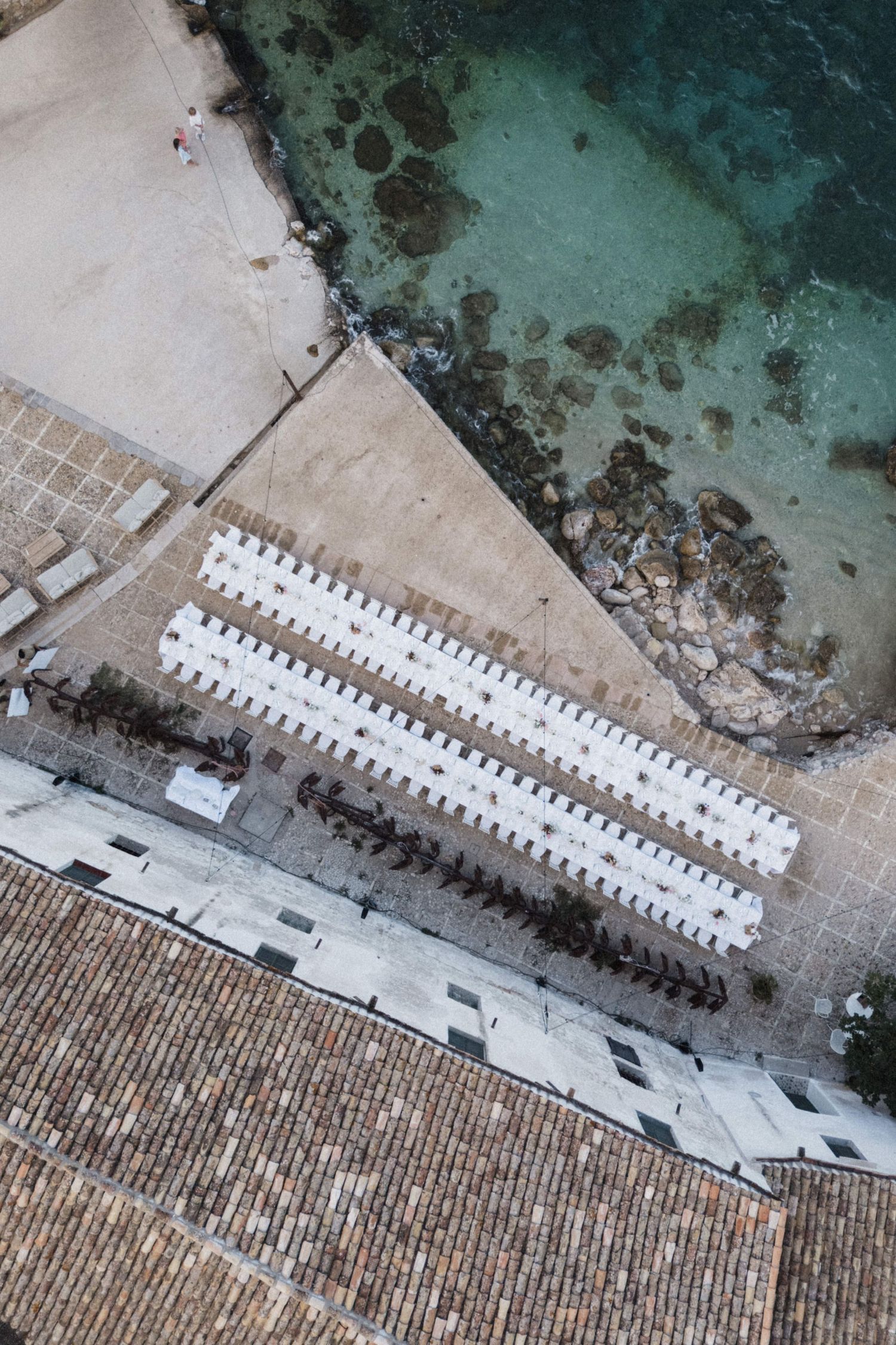 Top-down aerial view of white wedding reception tables arranged in rows beside turquoise coastal waters.