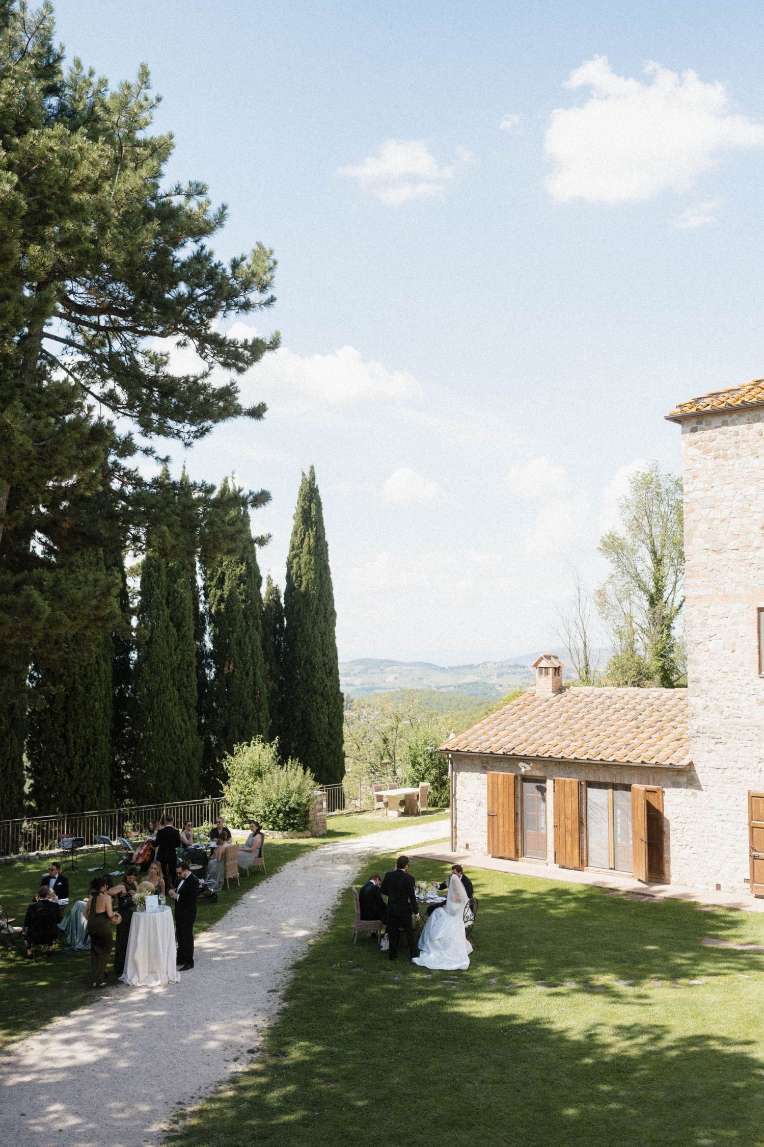 Wide shot of outdoor wedding guests gathered on a sunny lawn beside a rustic Tuscan stone building.
