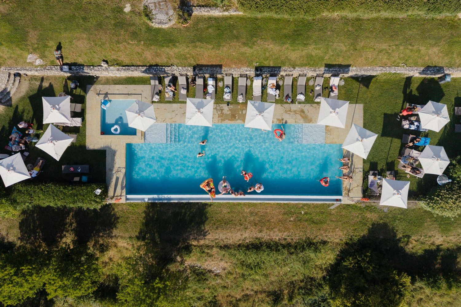 Aerial view of a rectangular outdoor swimming pool with white umbrellas and colorful floaties on a sunny day.