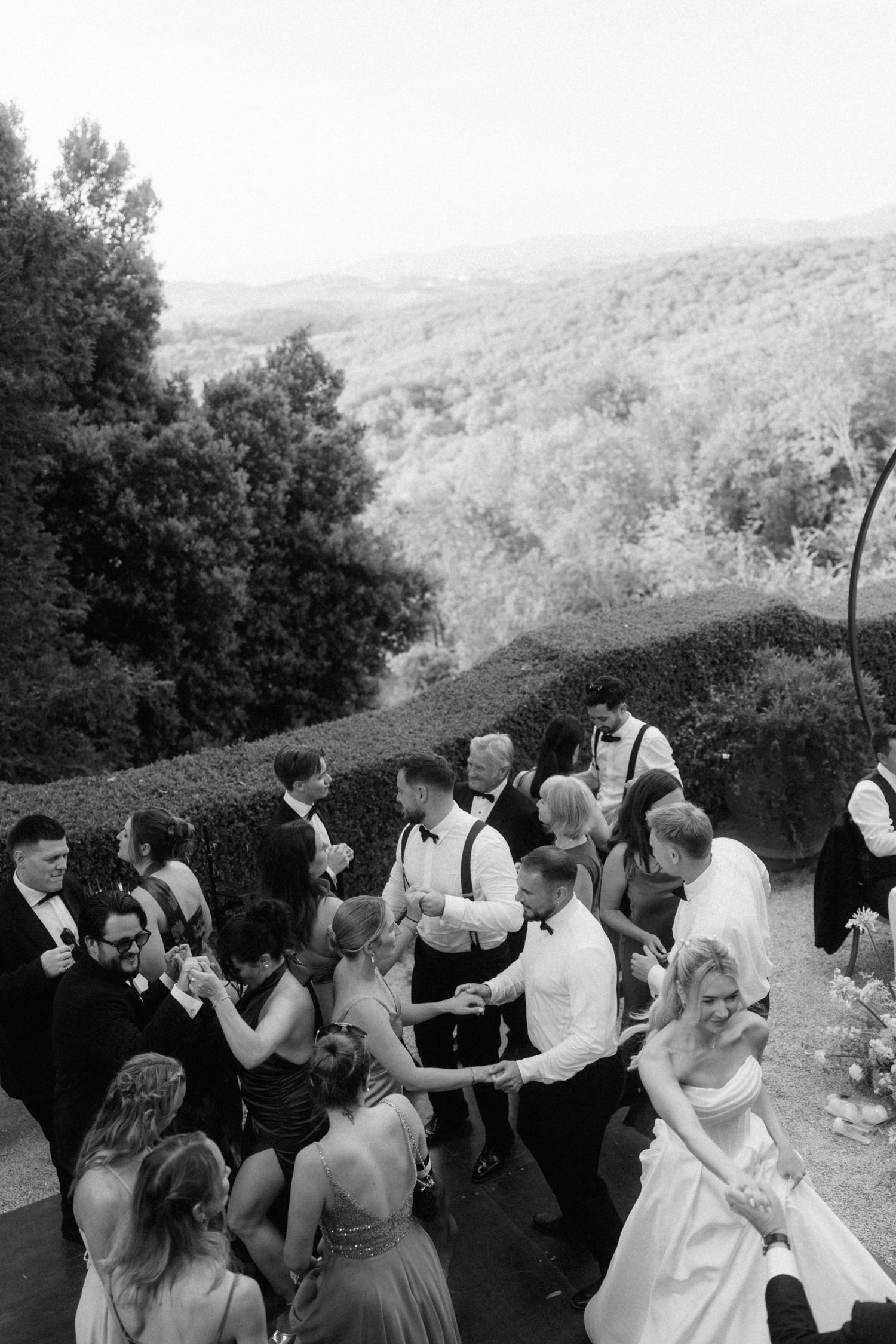 Aerial black and white view of wedding guests dancing outdoors with scenic hillside landscape backdrop.