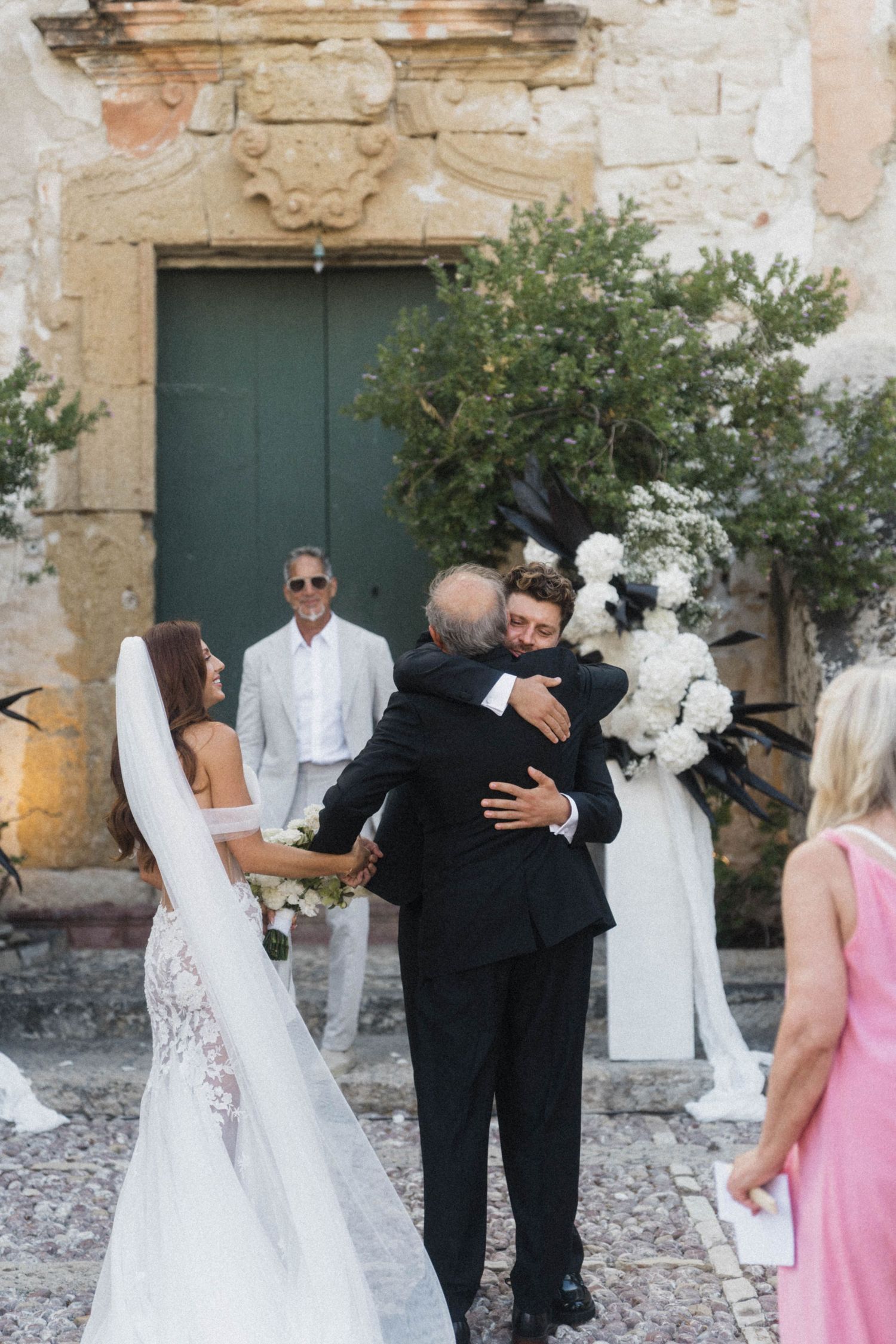 Groom embracing father of bride at outdoor wedding ceremony near floral arch.