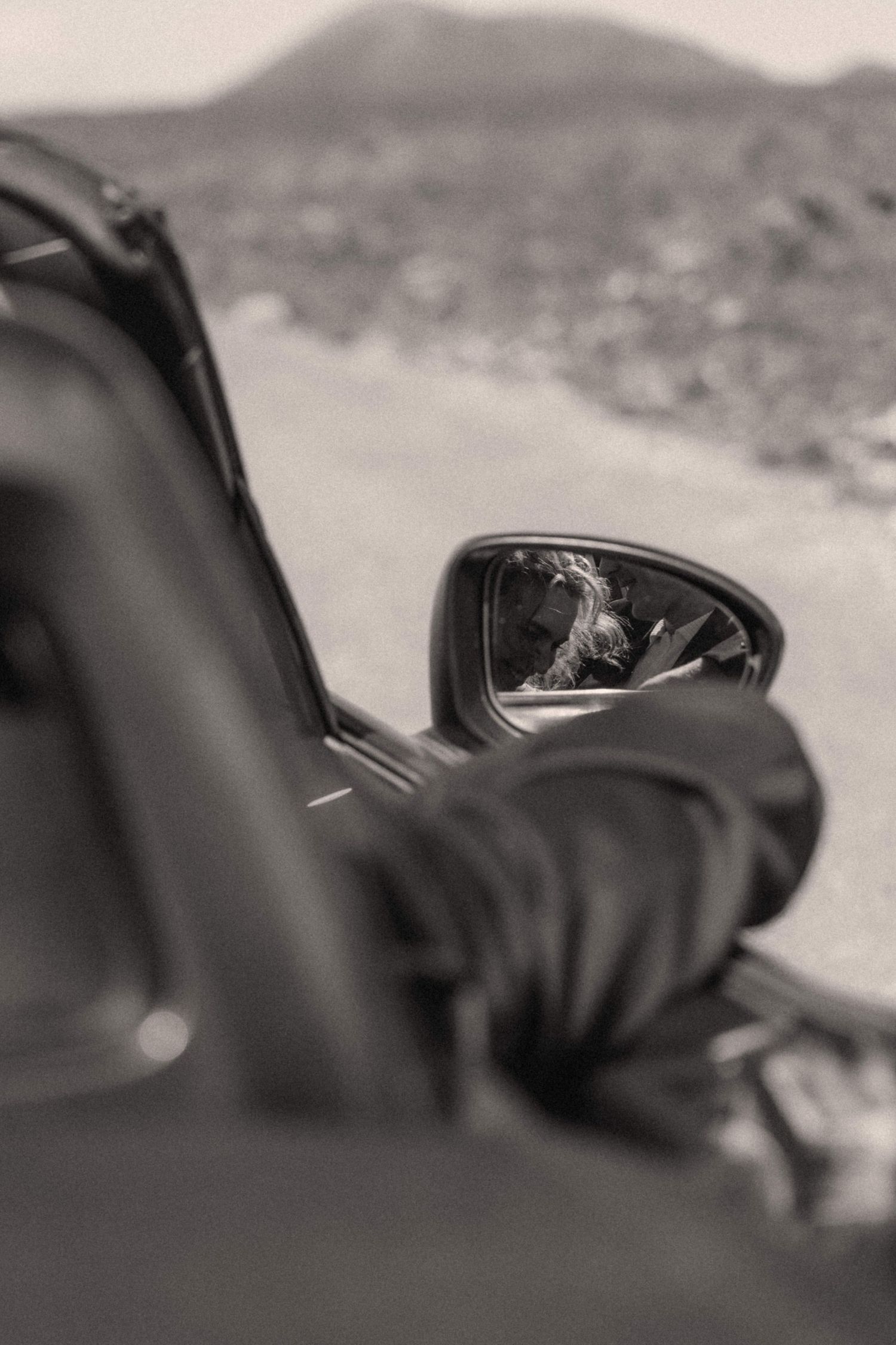 Black and white reflection of bride in vintage car side mirror on scenic road.