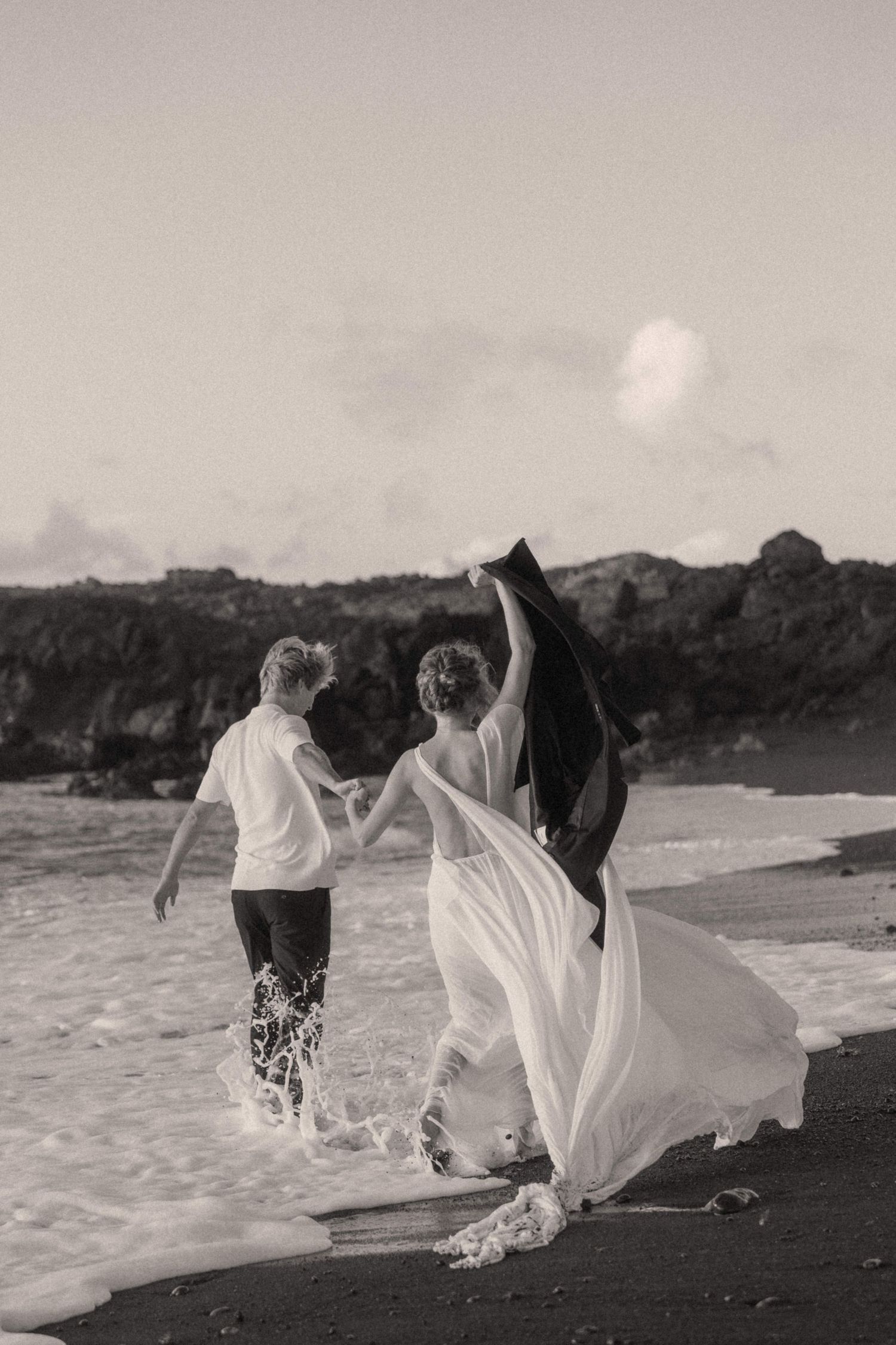 Black and white photo of bride and groom splashing through ocean waves on black sand beach.
