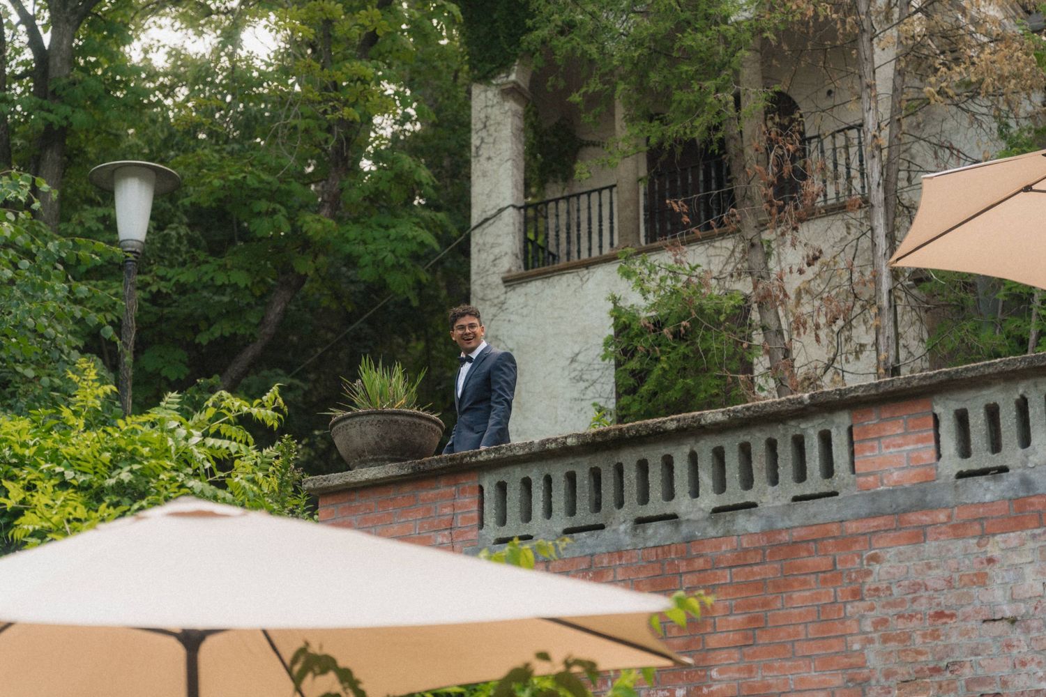 Elegantly dressed man standing on terrace overlooking lush garden venue with white umbrellas below.
