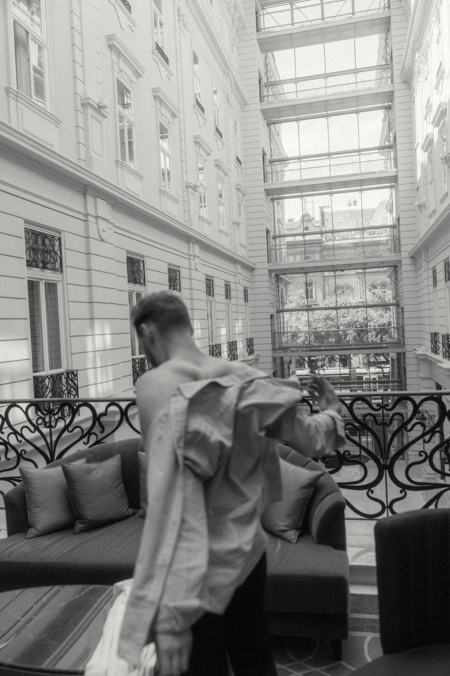 Black and white photo of a man in a suit jacket walking through an elegant hotel lobby atrium with glass ceiling.