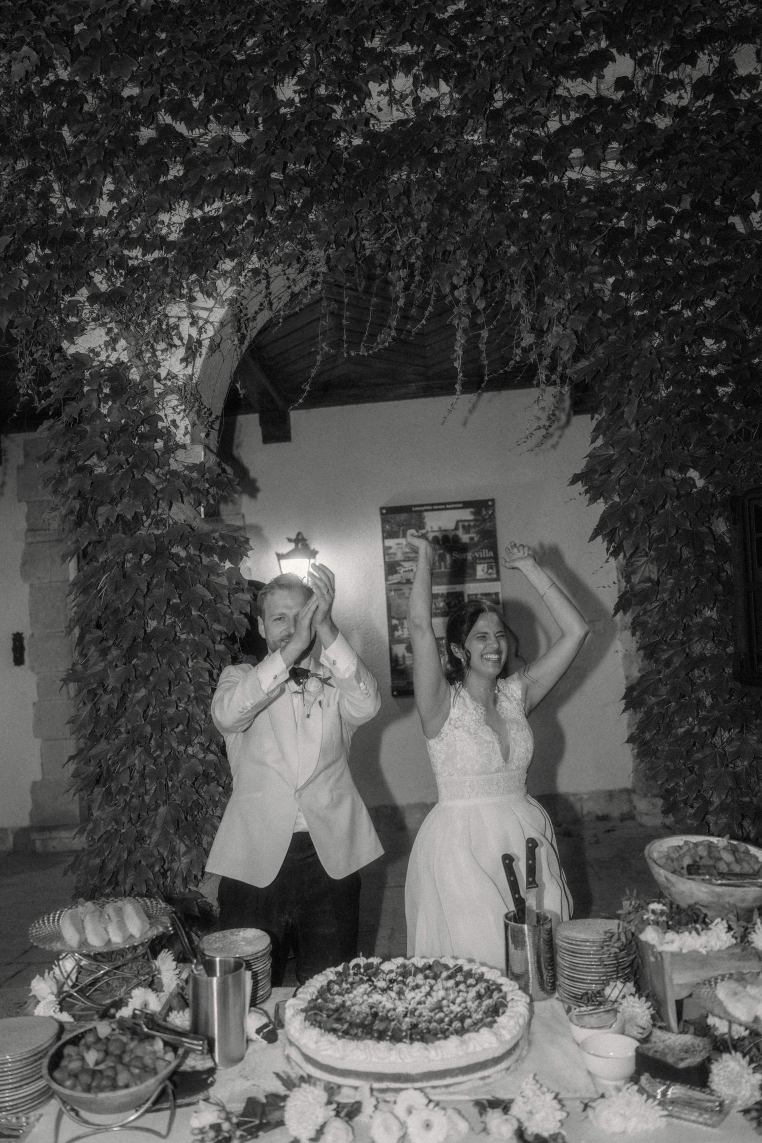 Black and white photo of a bride and groom celebrating at their wedding cake table.