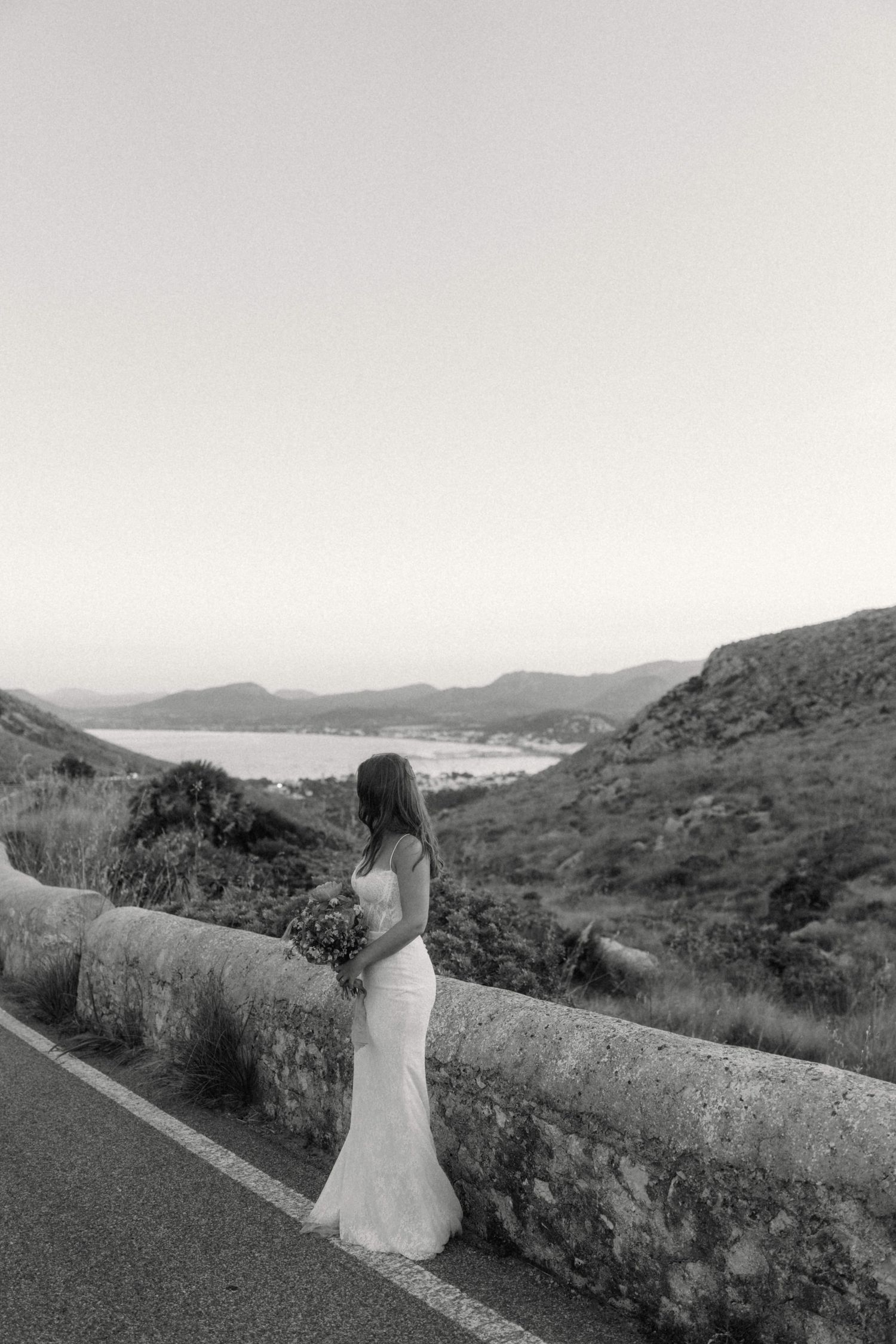 Black and white photo of a bride in a white gown holding a bouquet, standing by a stone road barrier with mountain views.