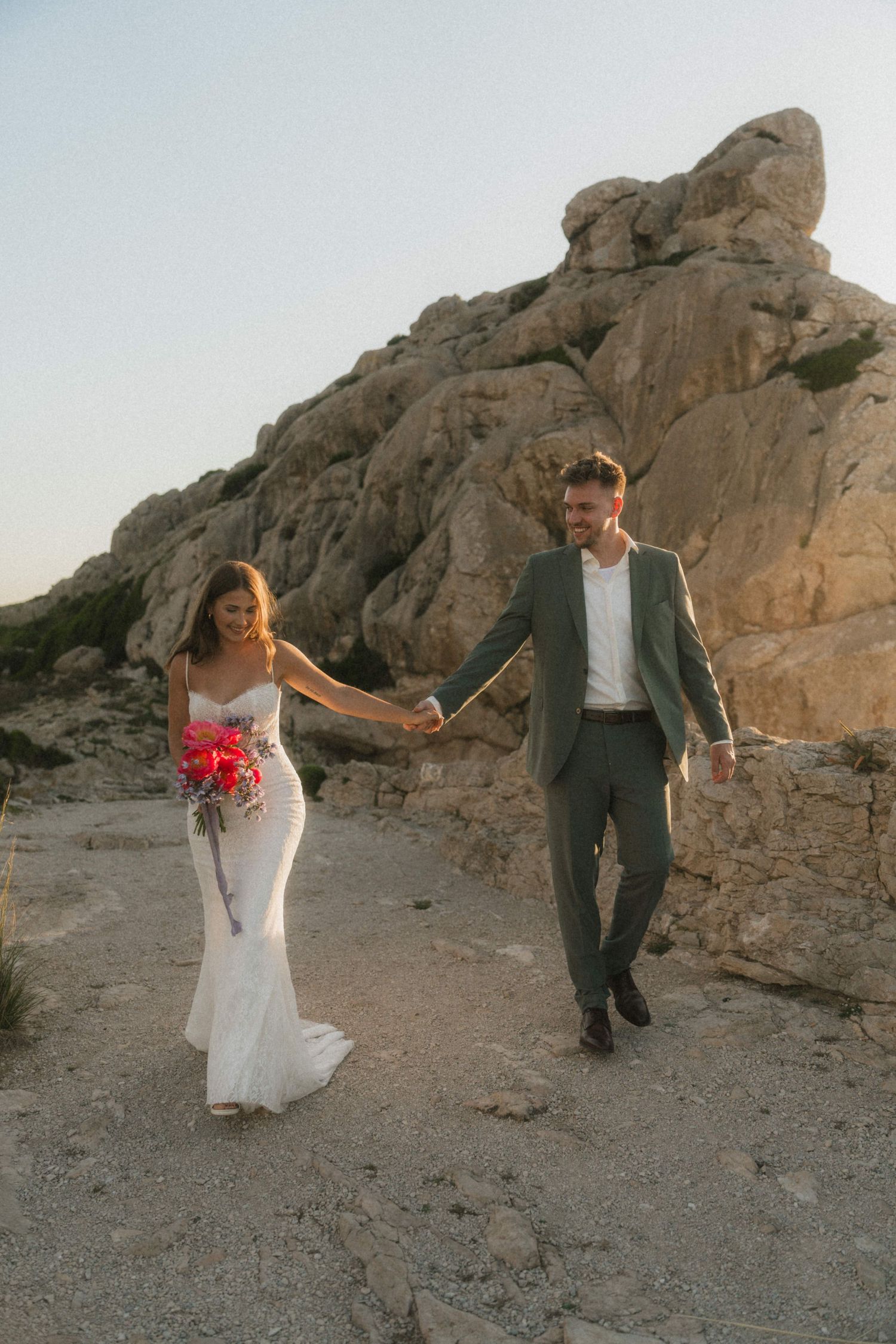 Bride and groom hold hands walking through rocky desert landscape at sunset, bride in lace gown holding red bouquet.