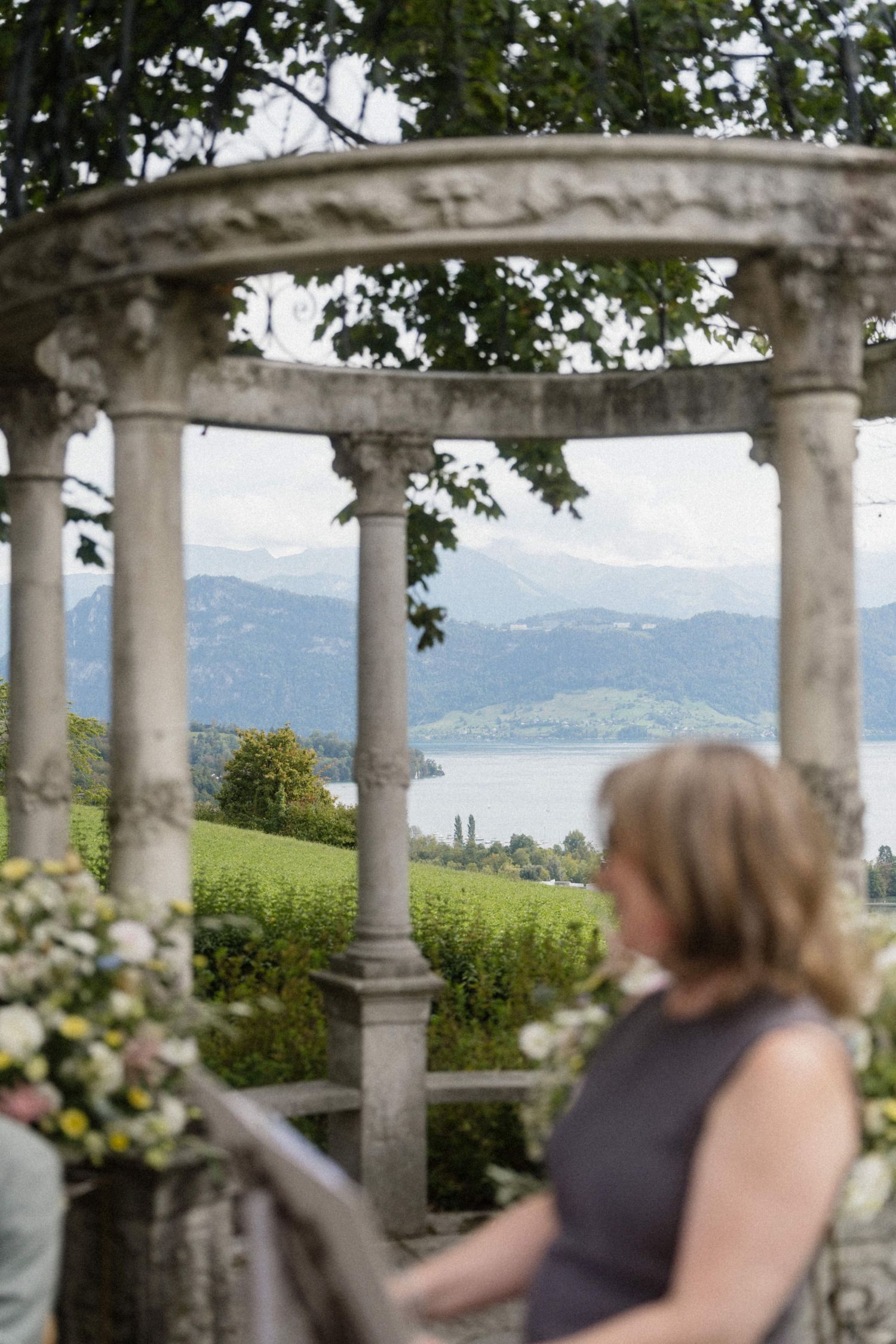 Stone pergola with elegant columns frames a scenic lake and mountain view, with a blurred woman in the foreground.